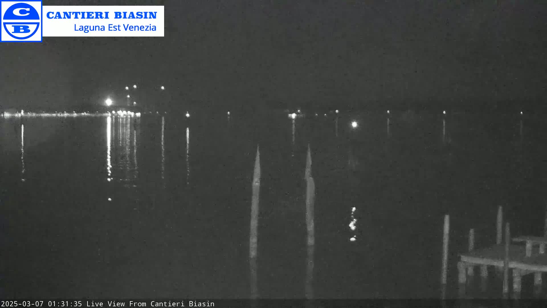 A nighttime view of a dark body of water with lights in the distance reflecting on the surface, and several posts in the water near a small dock in the foreground.