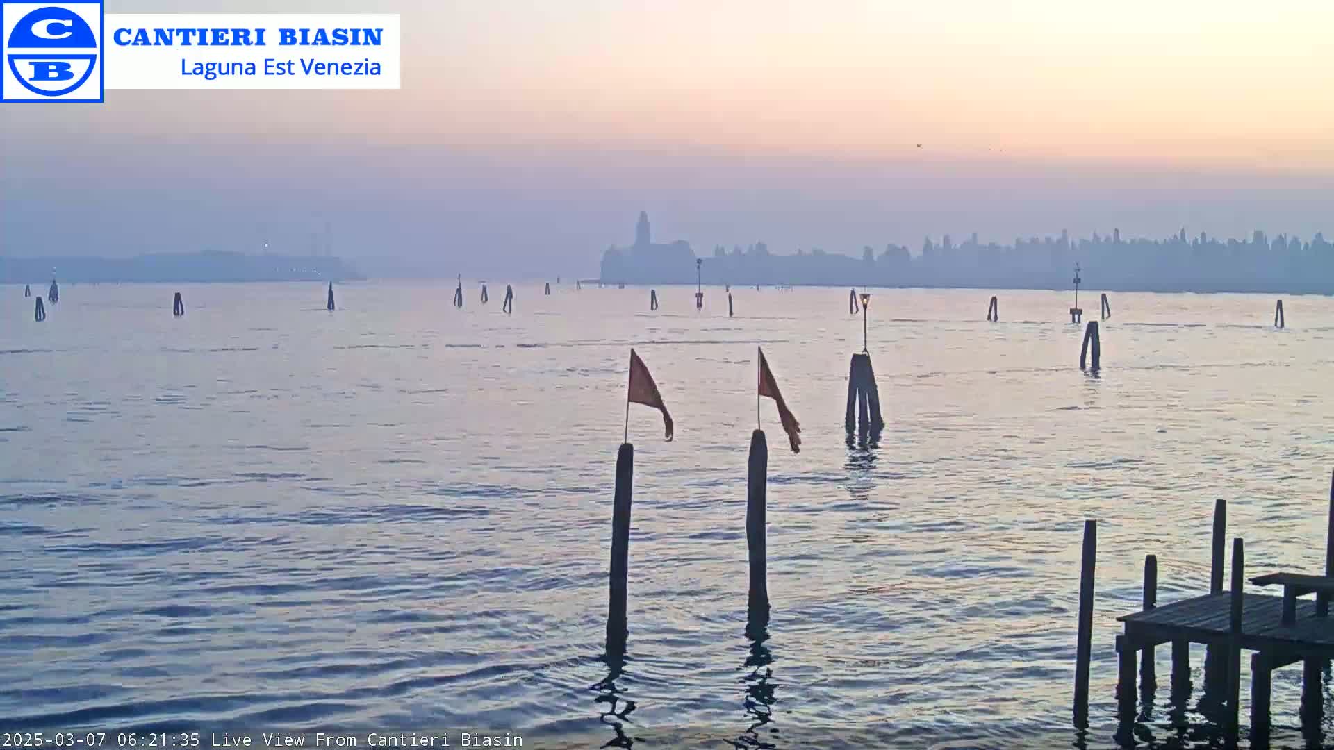 A hazy sunrise illuminates a calm body of water with numerous wooden posts and small orange flags, a distant hazy cityscape visible in the background.