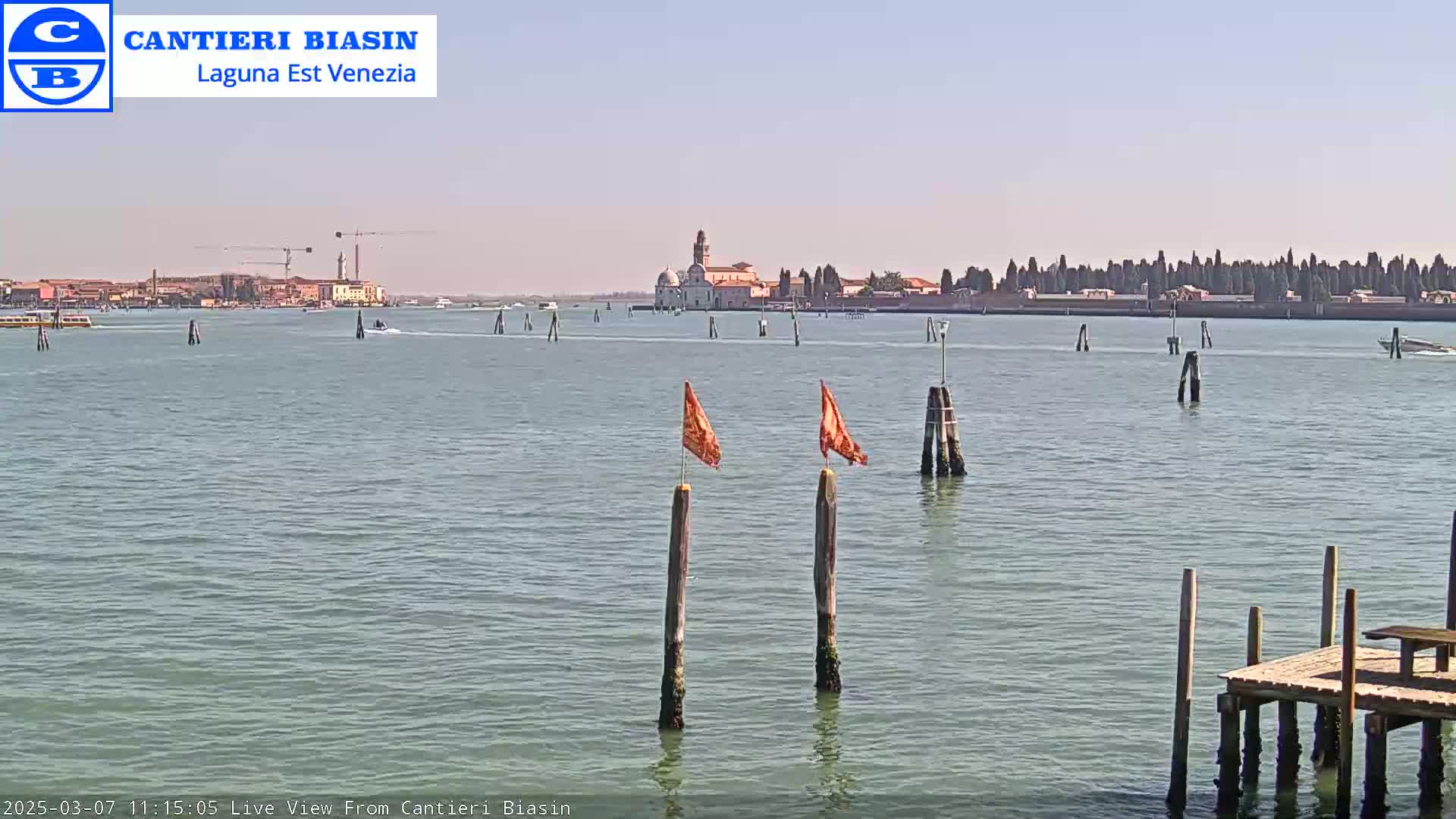 A calm body of water shows a distant island with buildings under a clear, sunny sky, with several wooden posts and orange flags in the foreground.