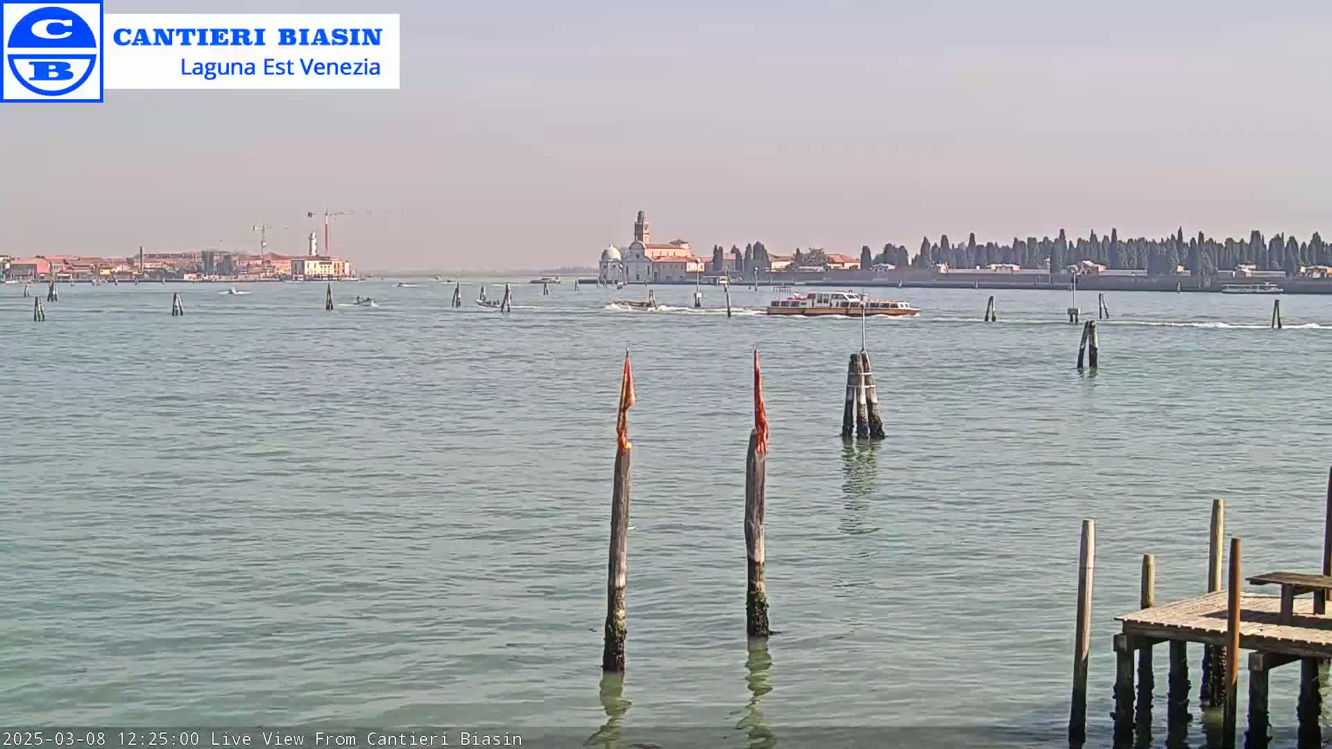 A calm body of water shows a distant island with buildings under a clear sky, featuring several boats and wooden posts in the foreground.