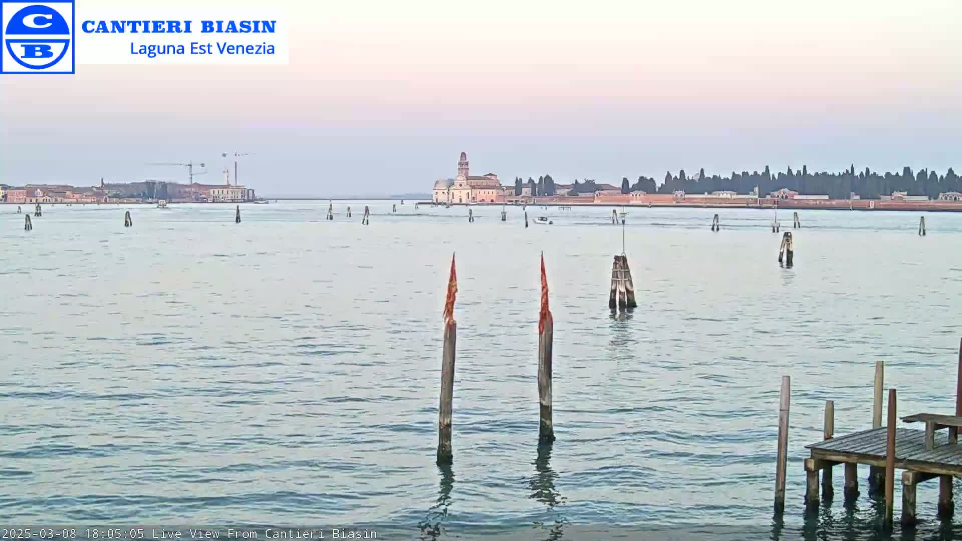 A calm body of water shows a distant island with buildings under a pale, pinkish sky.