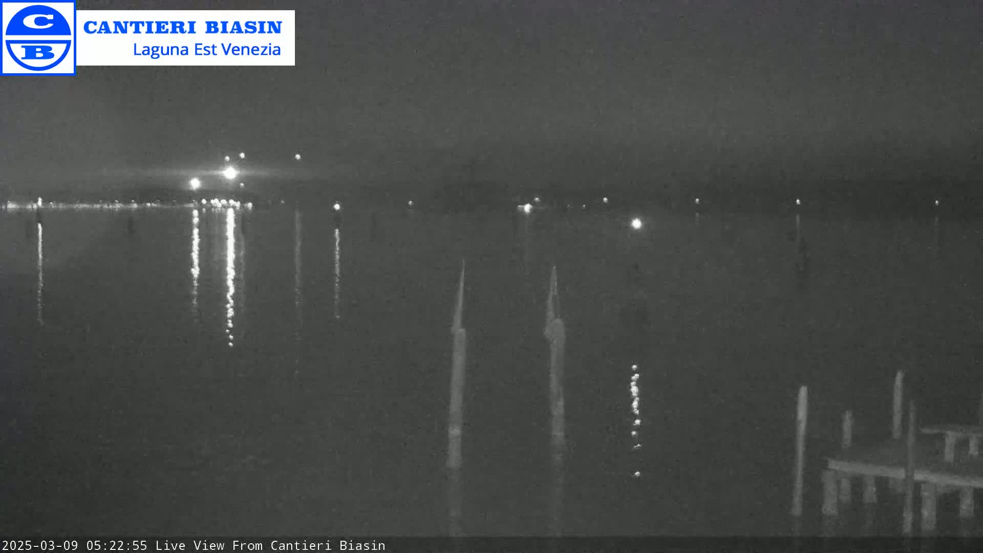 A nighttime view of a calm body of water with lights reflecting on the surface and several tall, thin posts in the water near a structure on the right.