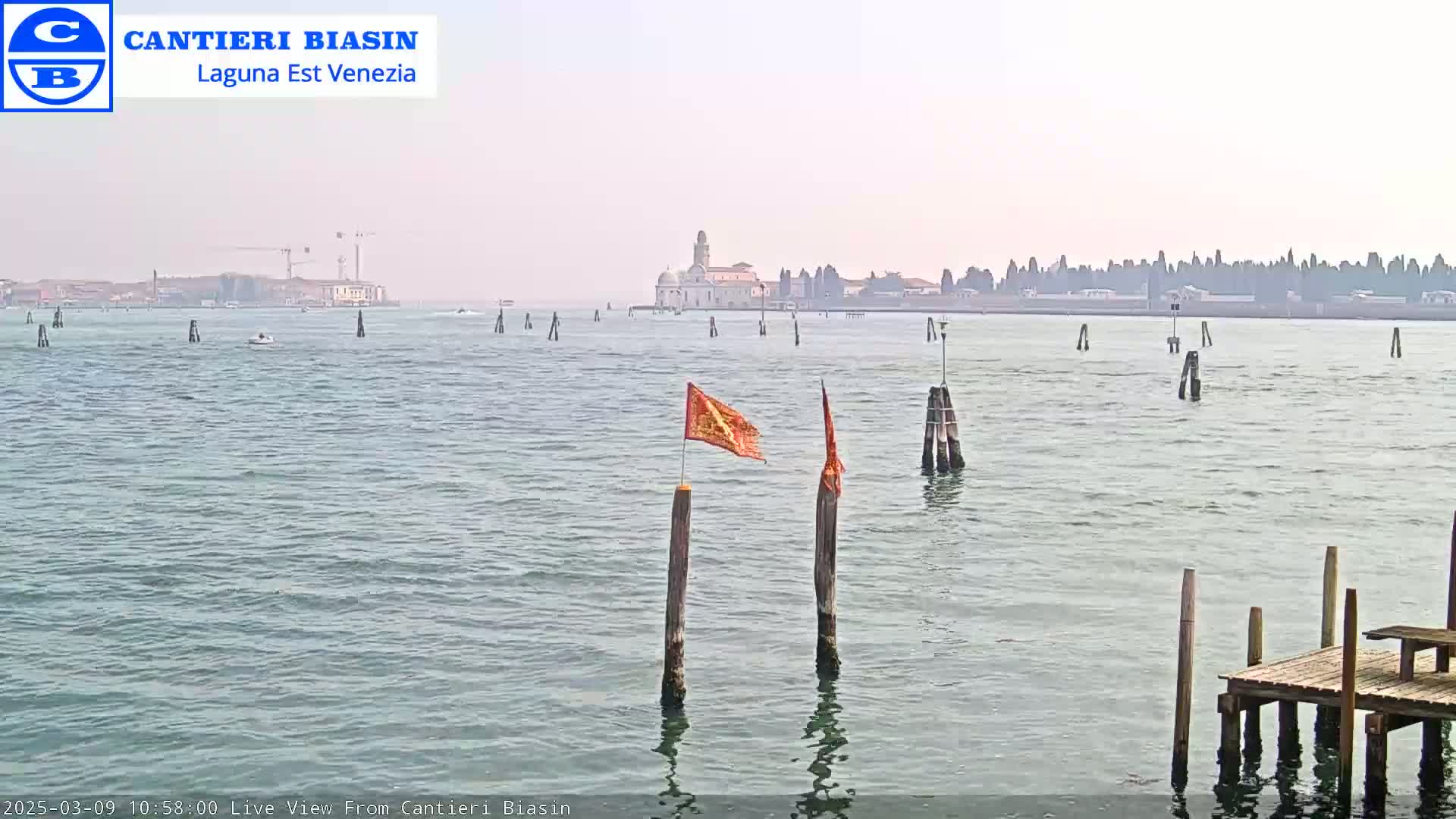 A hazy view across calm water shows a distant island with buildings, several wooden posts in the water, and a small orange flag on a post in the foreground.