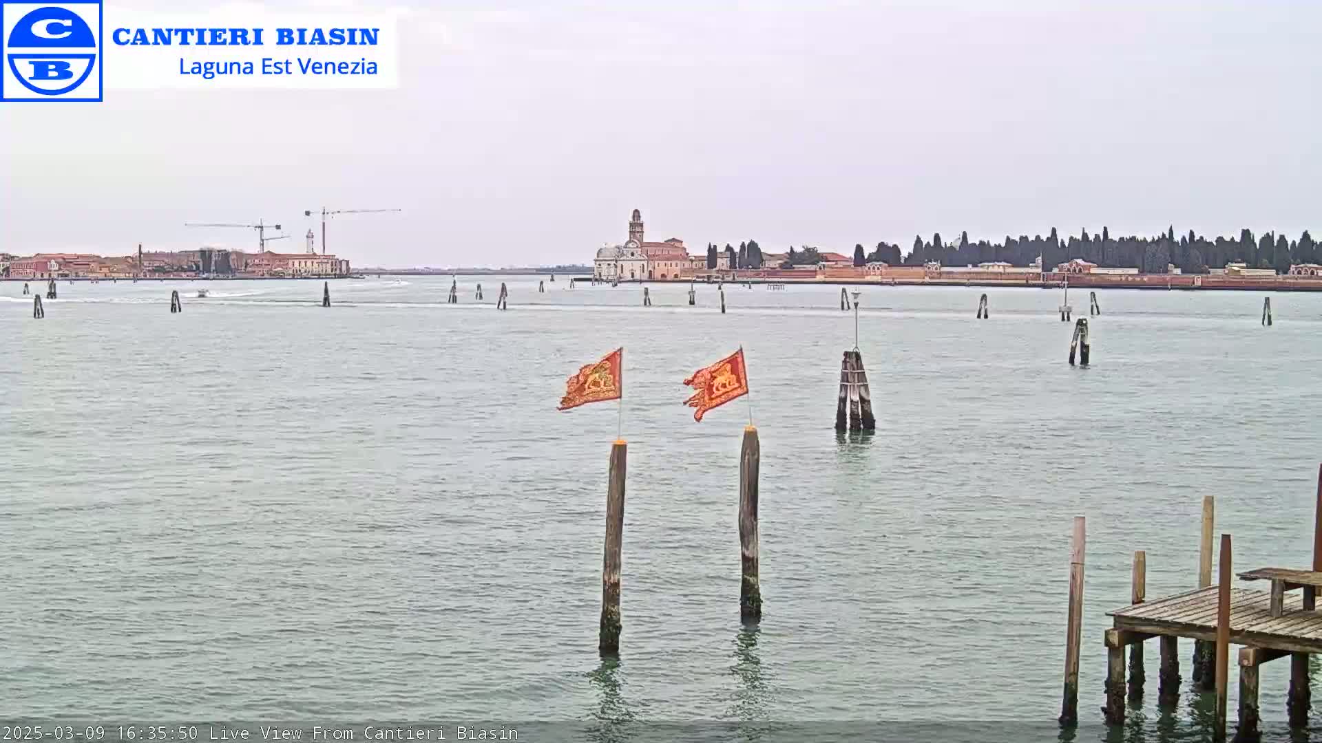 A calm body of water shows a distant island with buildings under an overcast sky, and two flags are mounted on wooden posts in the foreground near a small dock.