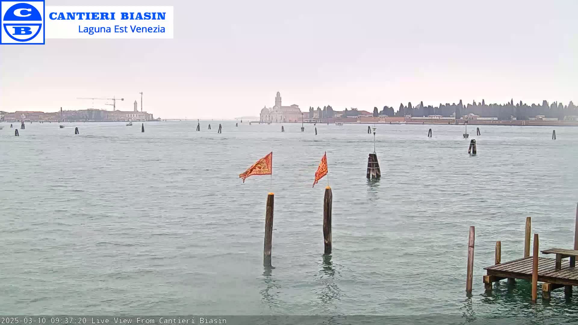 A calm body of water shows a distant cityscape under an overcast sky, with two orange flags on posts in the foreground and a small wooden dock.