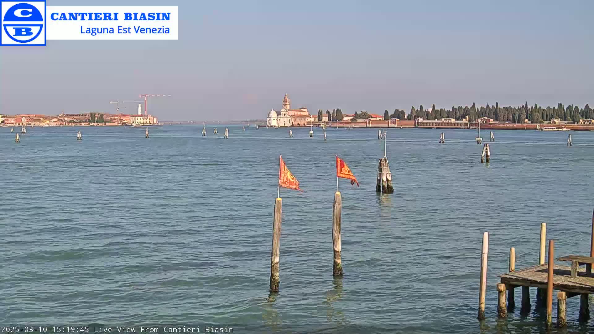 A calm body of water shows a distant island with buildings, a construction crane, and two orange flags on wooden posts in the foreground on a clear, sunny day.