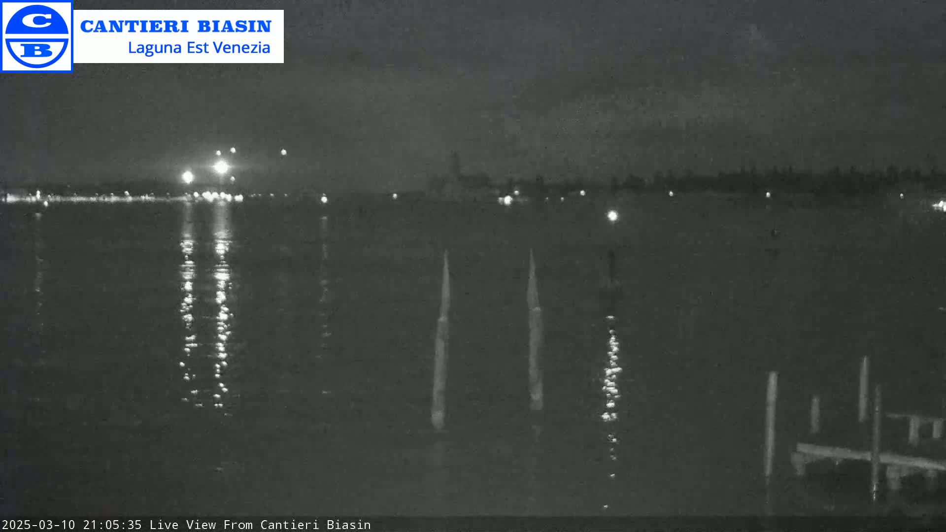 A nighttime view of a dark body of water with lights in the distance reflecting on the surface, and several thin posts visible in the foreground.