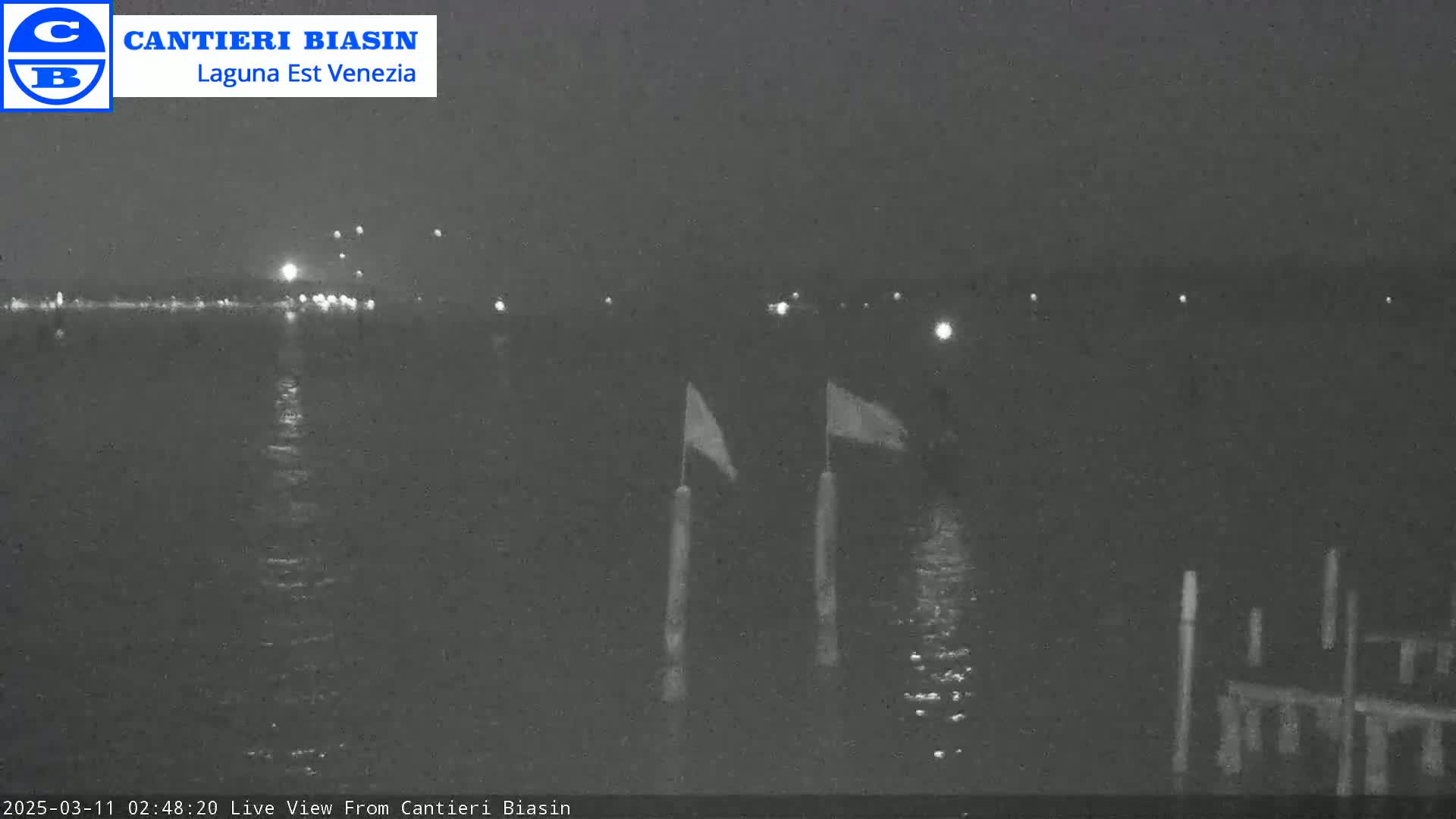 At night, two flags are visible on posts in dark water with a distant, faintly lit shoreline.