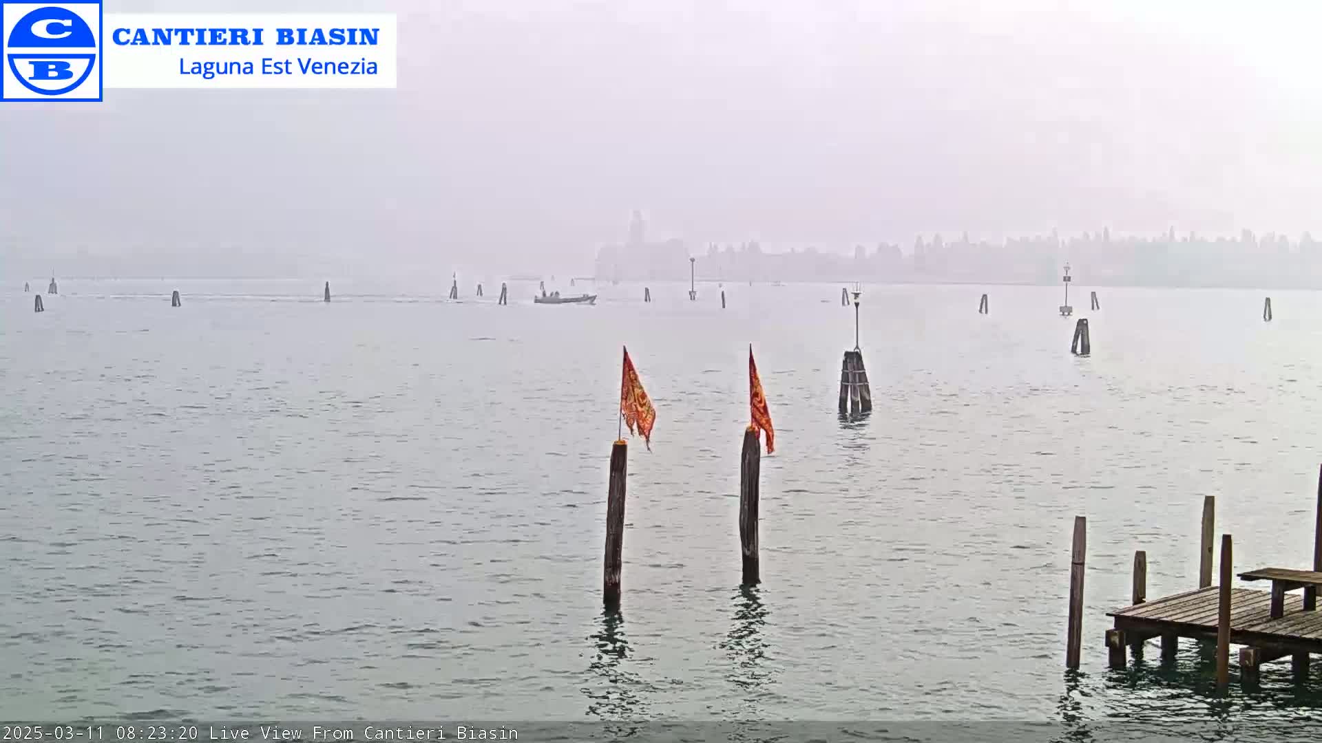 A foggy view of a calm body of water with several wooden posts and a small boat, featuring a distant hazy cityscape.
