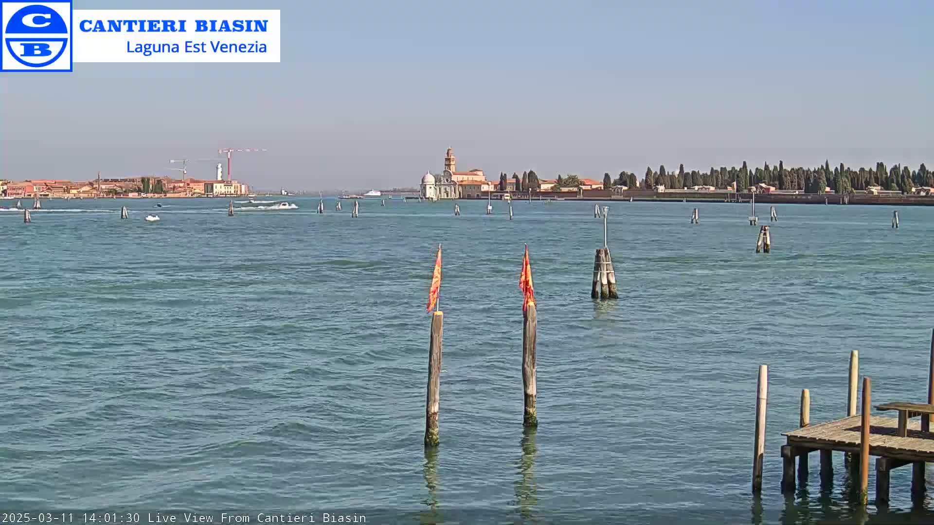A calm, sunny day on a body of water shows a distant island with buildings, a few boats, and two wooden posts with orange flags in the foreground.