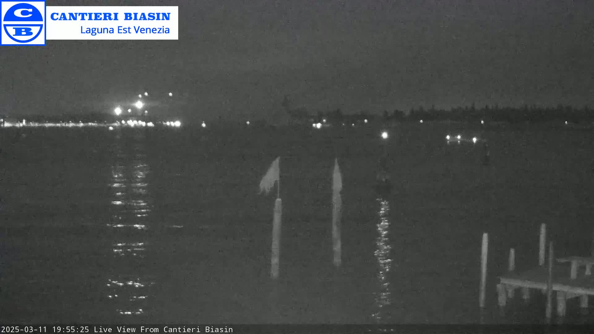 A nighttime view of calm water with distant lights on the shore and two posts with flags in the foreground.