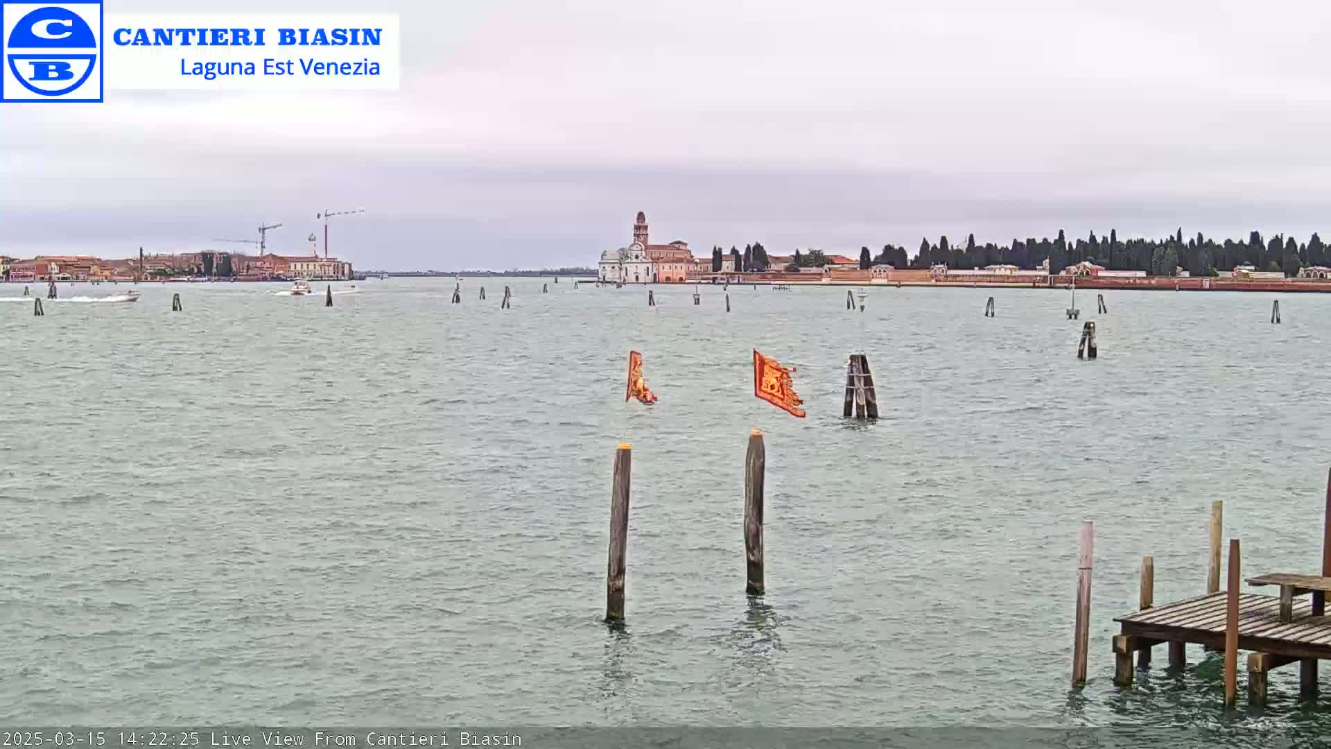 A calm body of water shows a distant island with buildings, trees, and a construction crane under an overcast sky, with several wooden posts and tattered orange flags in the water near a small wooden dock.