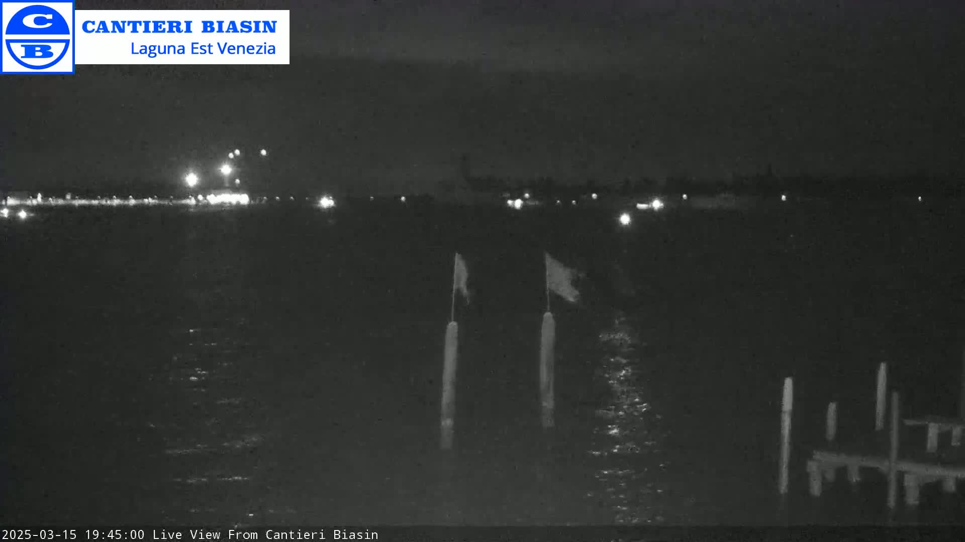 A nighttime view of dark water with a distant shoreline showing faint lights, two flagpoles with flags, and a dock in the foreground.