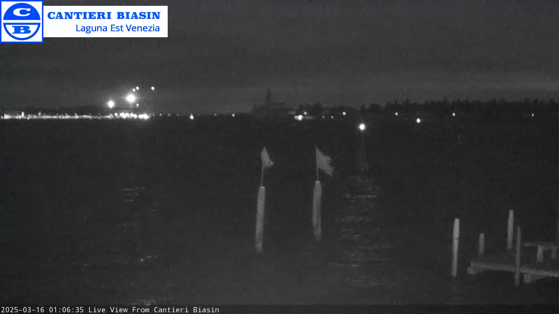 A nighttime, low-light view shows a dark body of water with a distant shoreline of lights, two flags on poles in the foreground, and a small dock.