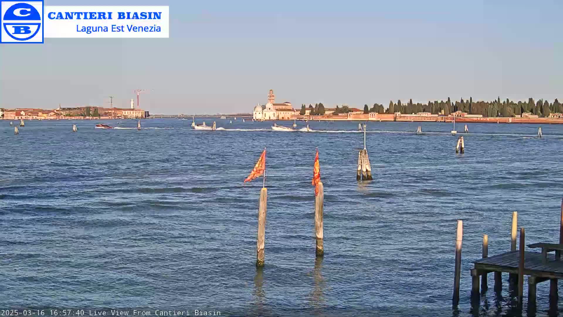 A calm body of water shows a distant island with buildings under a clear sky, and small boats are visible on the water near a small dock with orange flags on posts.