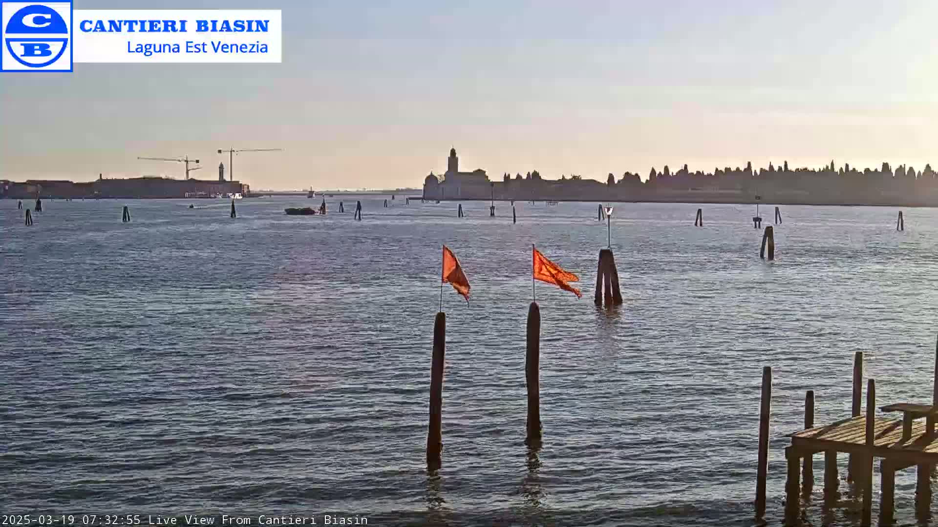 A calm body of water shows a distant city skyline under a clear sky, with orange flags marking wooden posts in the foreground and a small wooden dock in the bottom right corner.