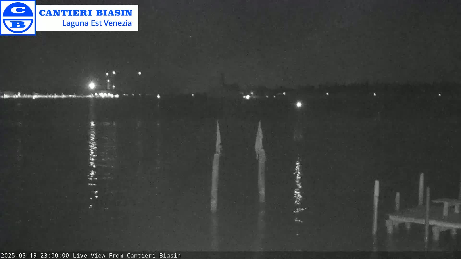 A nighttime view of calm water reflecting distant lights, with two tall, thin posts in the foreground and a dock to the right.