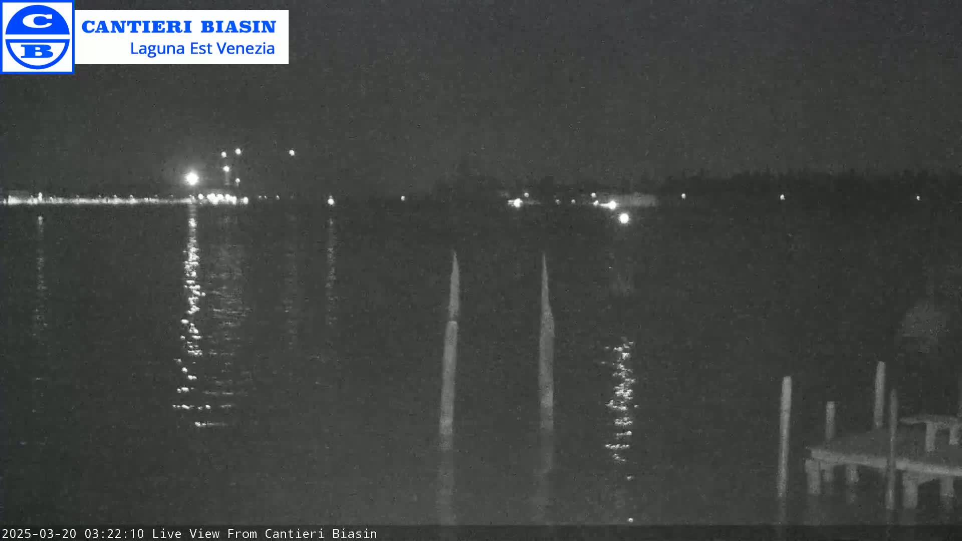 A nighttime view of a calm body of water with lights reflecting on the surface, showing a distant shoreline with lights and a dock in the foreground.