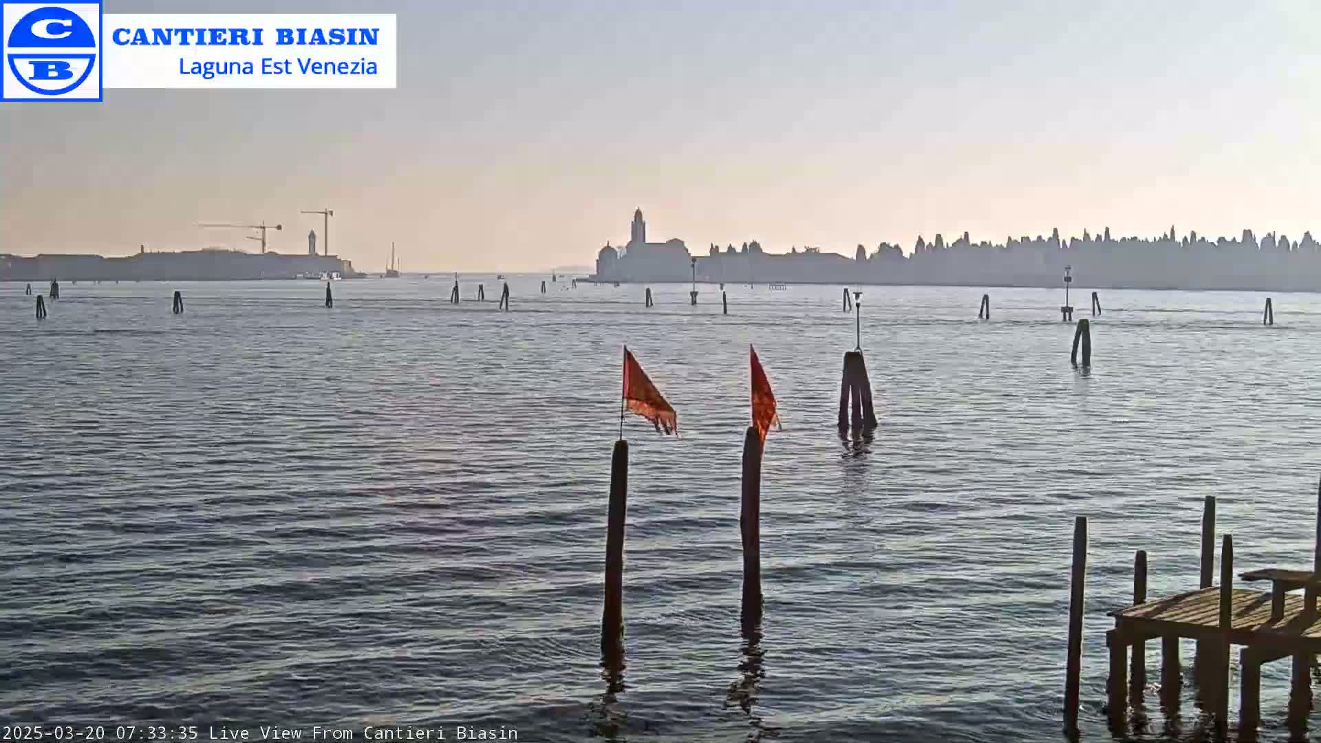 A calm body of water shows a distant city skyline under a clear, light sky with a few orange flags on wooden posts in the foreground and a small wooden dock to the right.