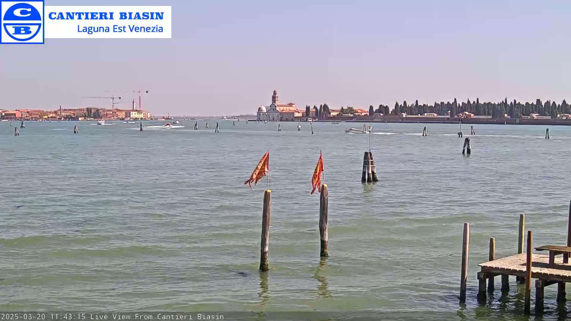 A calm, sunny day shows a body of water with a distant island featuring buildings and trees, and a closer view of wooden posts with flags and a small dock.