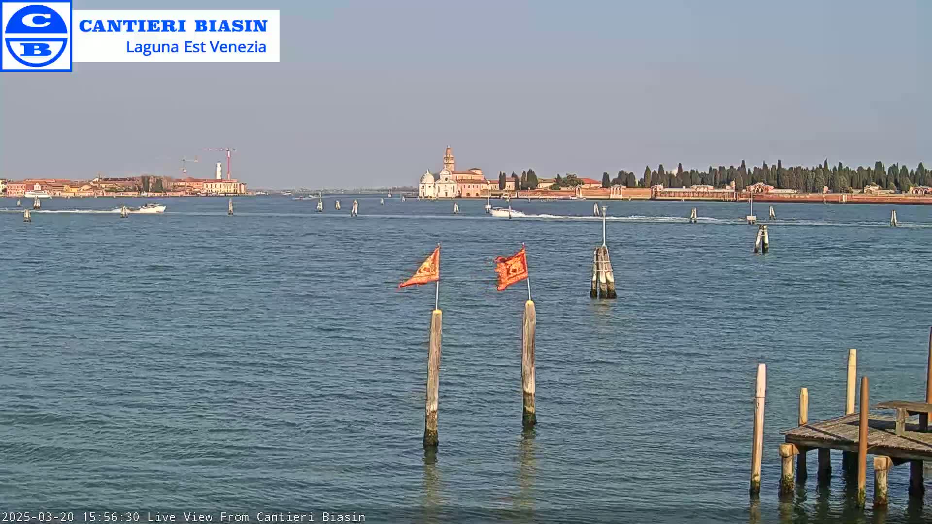 A calm body of water shows a distant island with buildings, a few boats, and two orange flags on posts in the foreground.