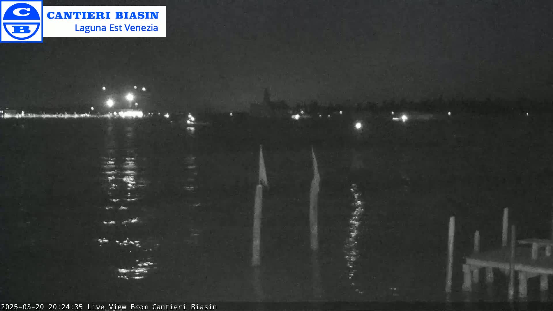 A nighttime view across a dark body of water shows a distant shoreline with faint lights, two upright posts in the foreground, and a small dock to the lower right.