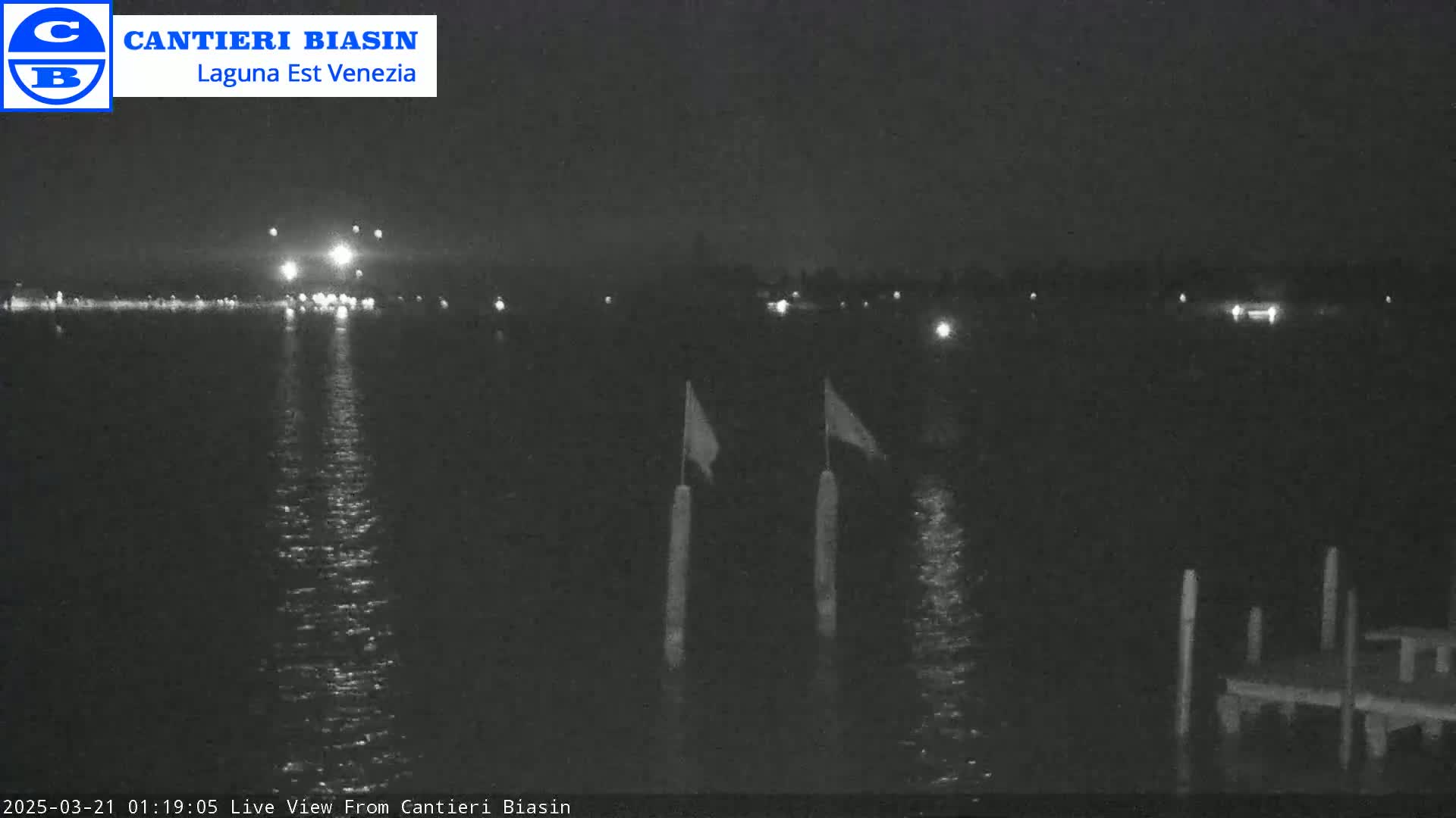 A nighttime view of calm water reflecting lights from a distant shoreline, with two thin posts and flags visible in the foreground and a small dock to the right.