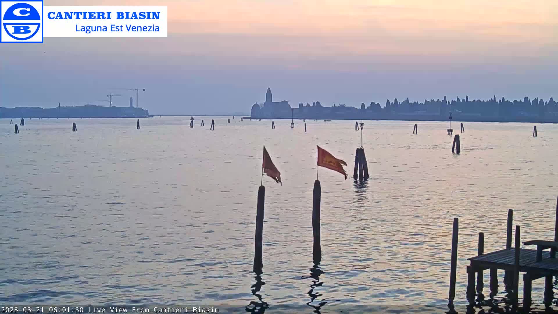 A calm body of water with wooden posts and small flags, showcasing a distant cityscape under a soft, pinkish-purple sunrise.
