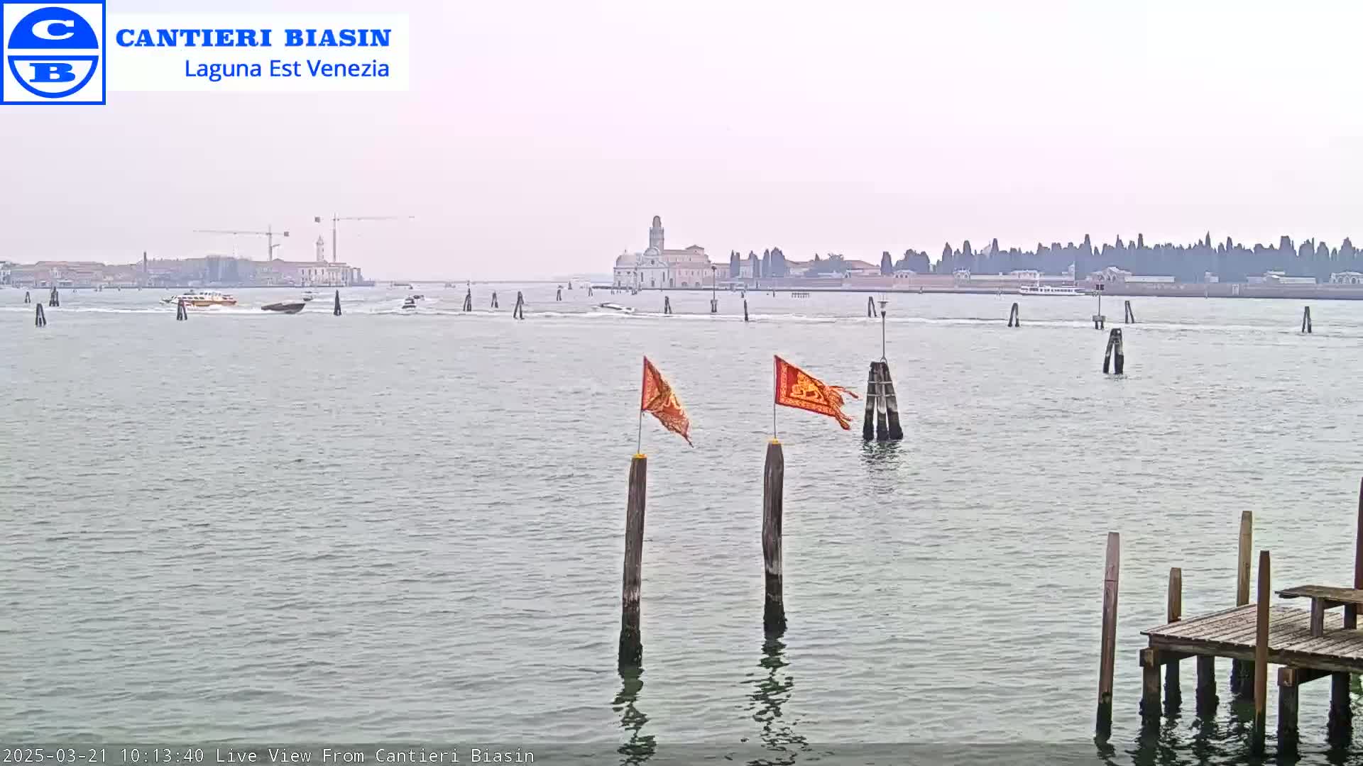 A calm, overcast day reveals a Venetian waterway with several boats, a distant island featuring buildings and a church, and two orange flags flapping atop wooden posts near a small dock.