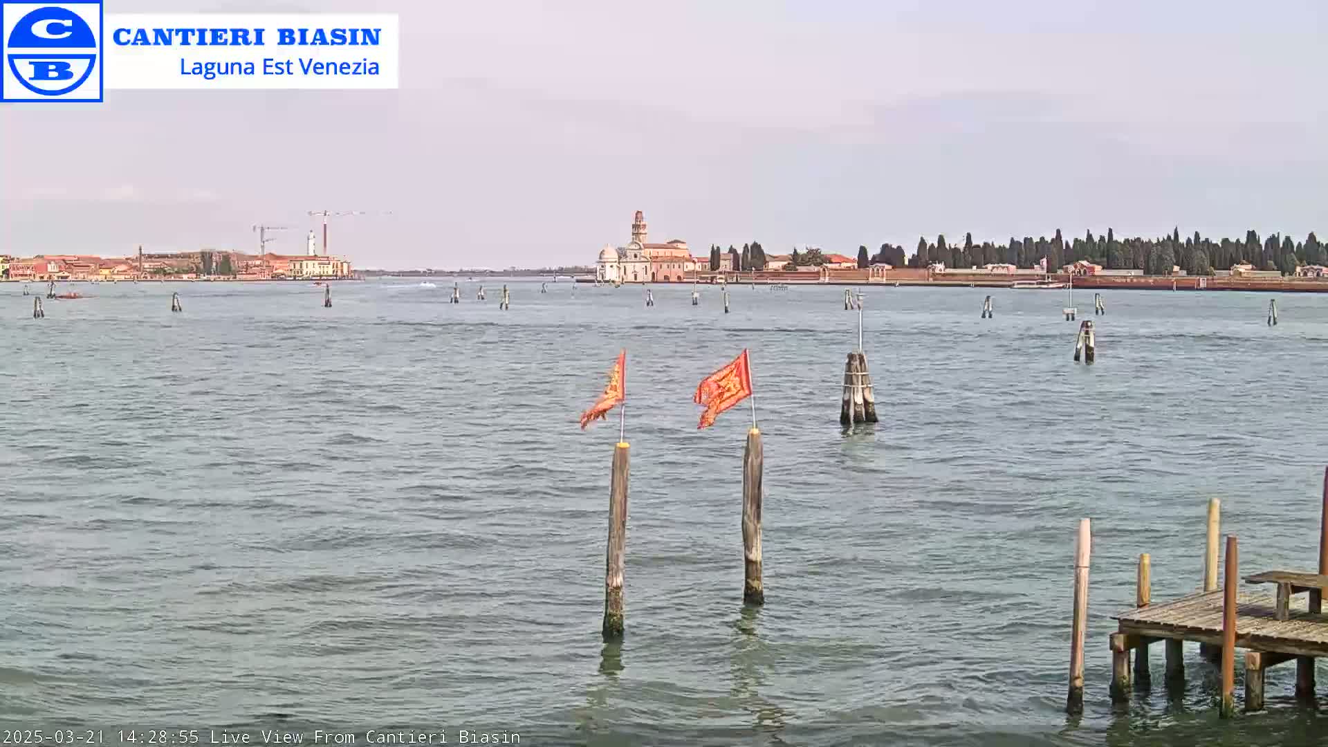 A calm body of water shows a distant island with buildings and trees under a hazy sky, with two orange flags on posts in the foreground and a small wooden dock to the right.