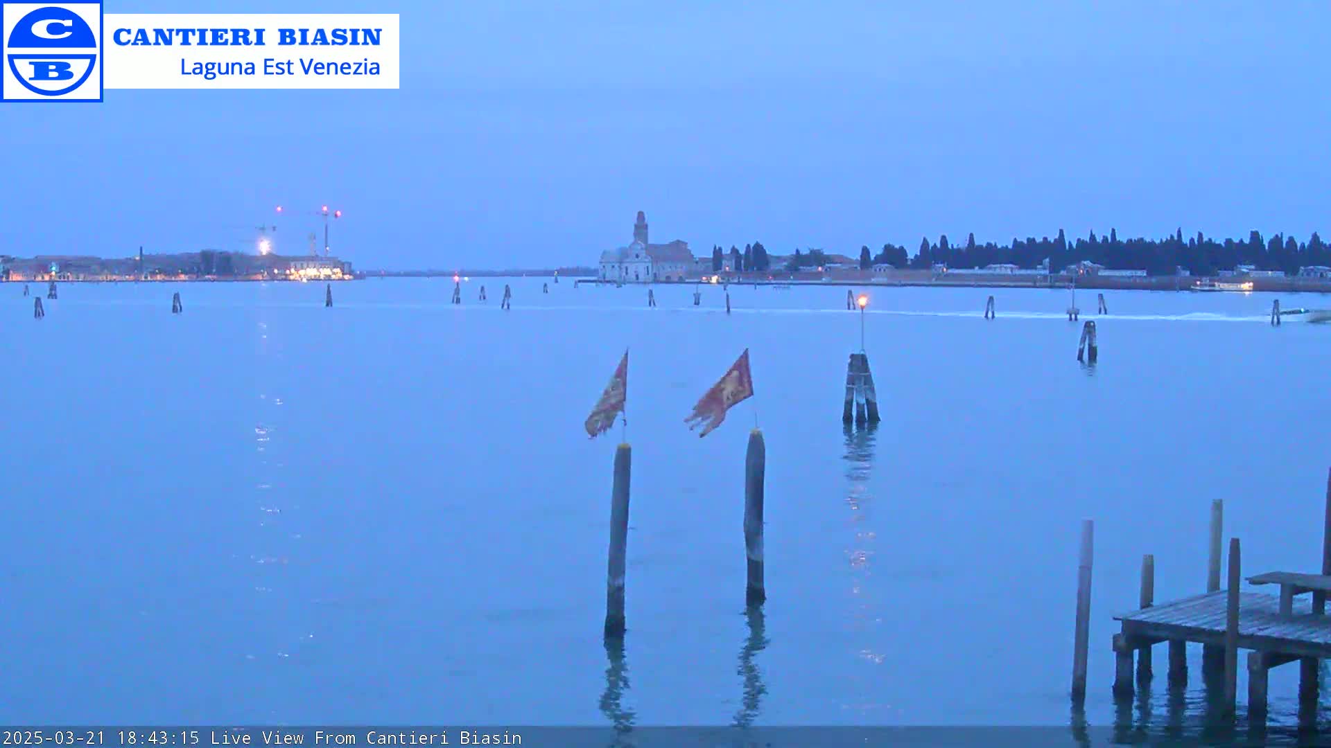 A tranquil, twilight view of a waterway shows a distant island with buildings, construction cranes, and numerous markers extending into the water; two flags are mounted on posts in the foreground, near a small wooden dock.