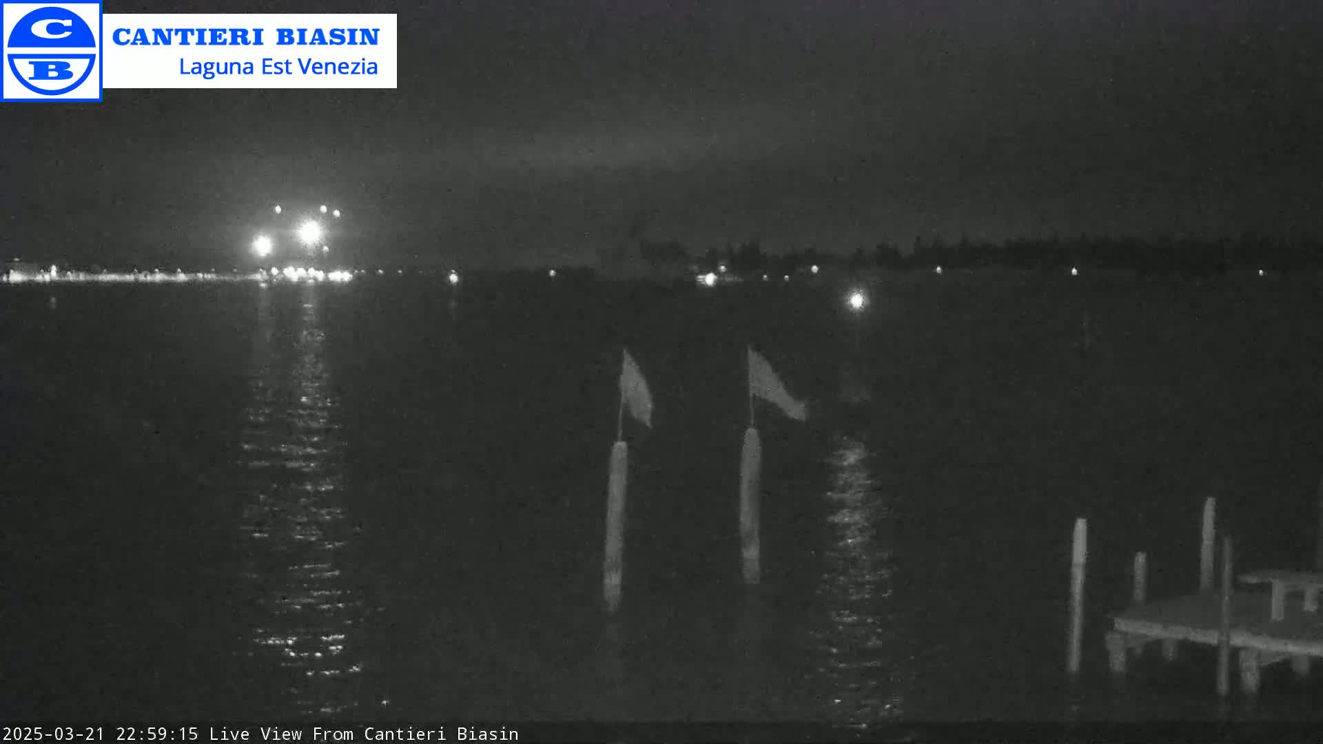 A nighttime view of a calm body of water with a distant shoreline illuminated by lights, two flags on poles, and a small dock in the foreground.