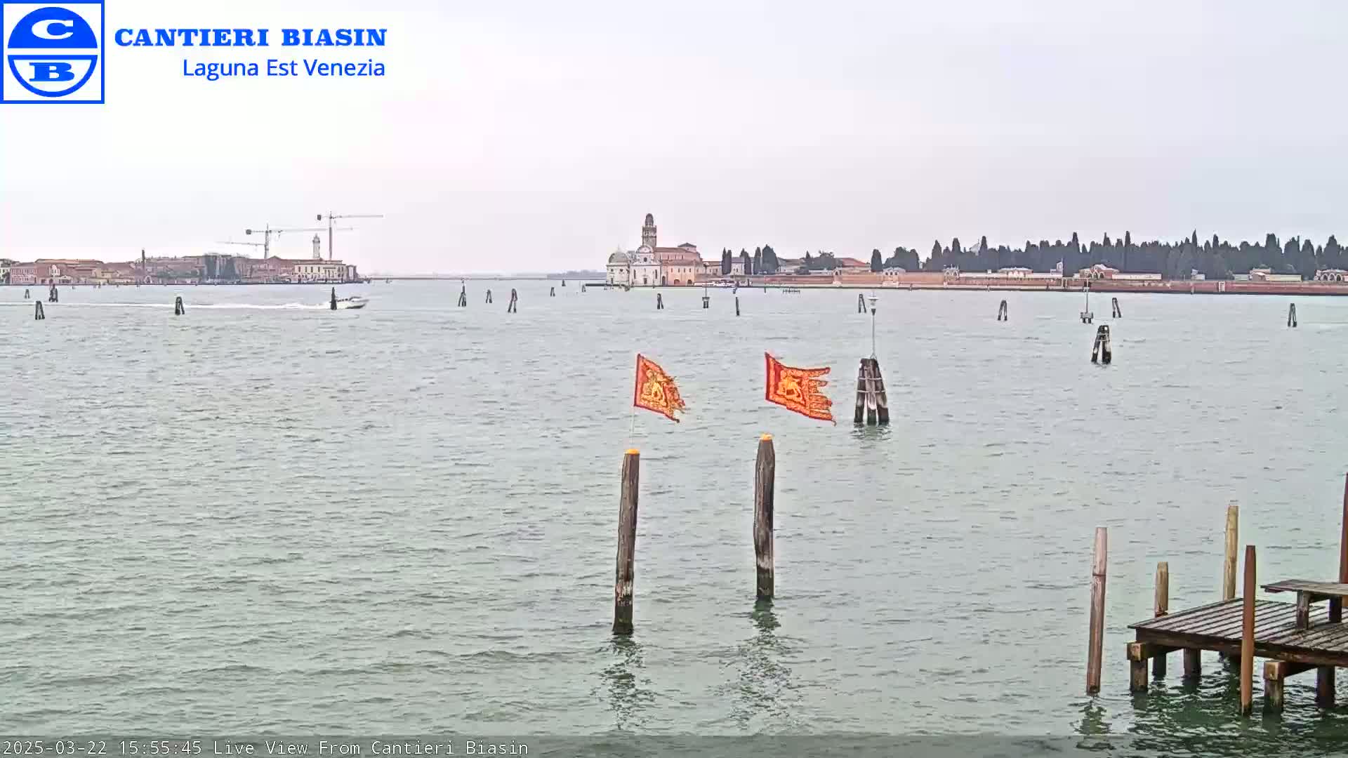 A calm body of water shows a distant island with buildings and construction cranes under an overcast sky, with two orange flags on posts in the foreground and a small dock on the right.