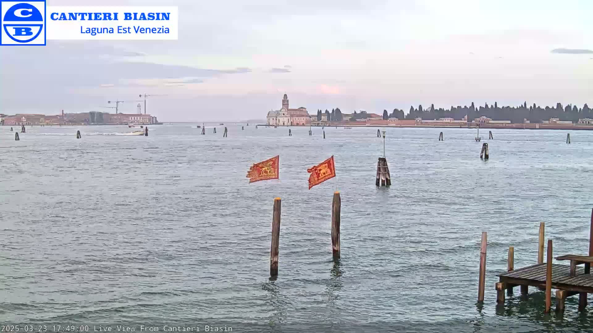 A calm, overcast day shows a waterway with a distant island featuring buildings and trees, two tattered flags on posts in the foreground, and a small dock to the lower right.