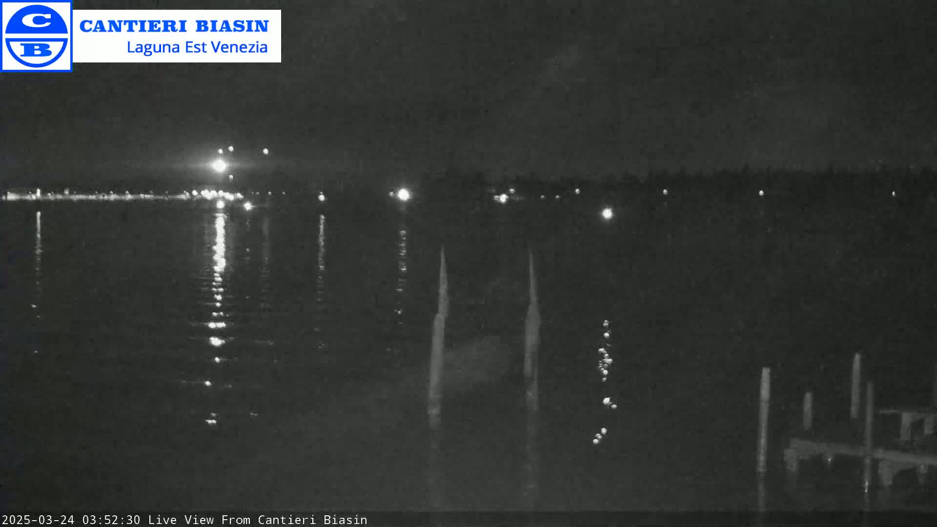 A nighttime view of a calm body of water with lights reflecting on its surface, and a dark shoreline with sparsely placed lights in the distance.