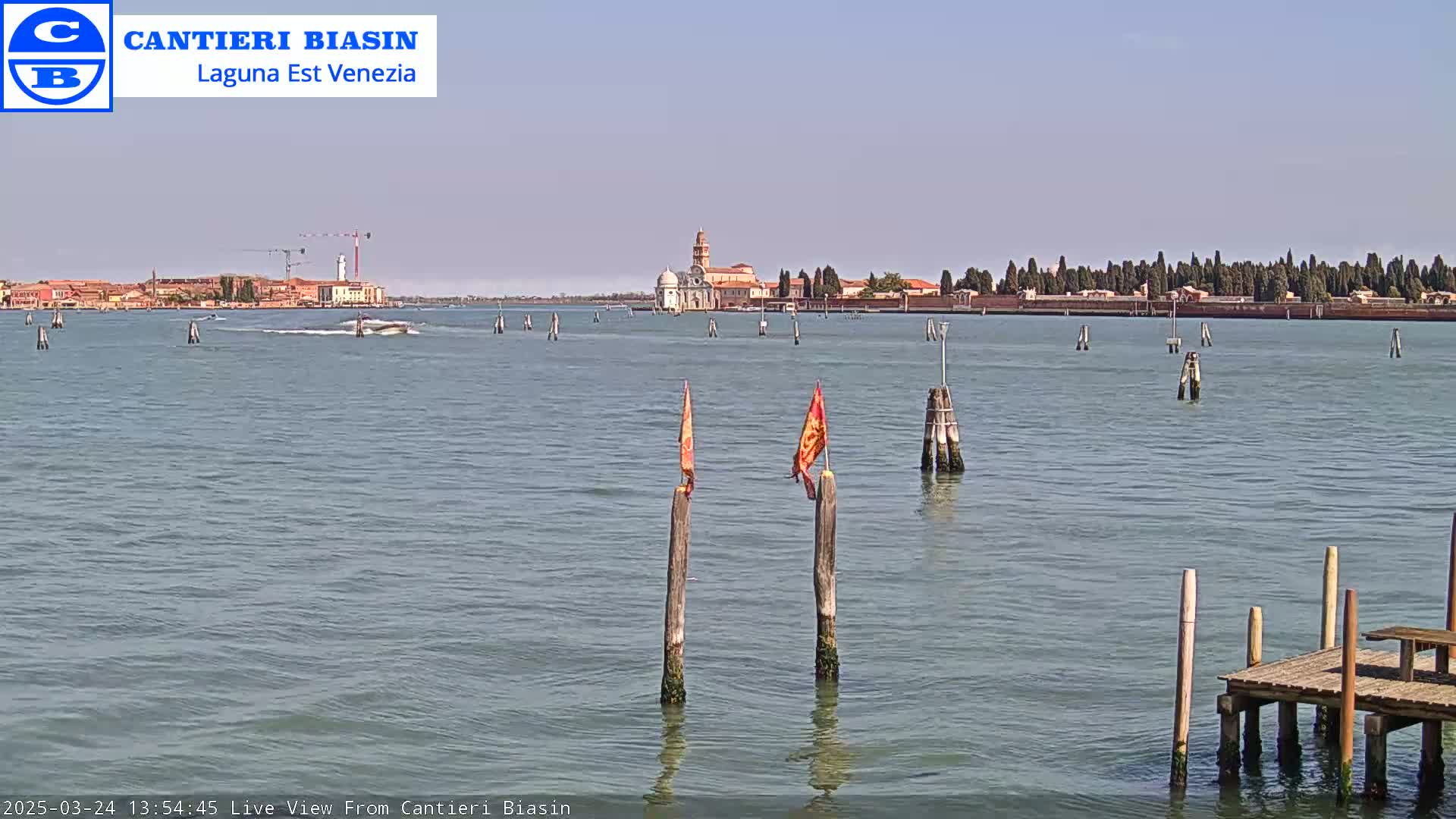 A calm, sunny day shows a Venetian island with buildings and trees across a body of water, marked by wooden posts and small boats.