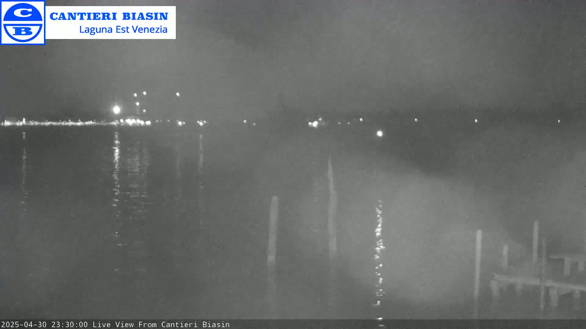 A nighttime view of a calm body of water shows distant lights on the opposite shore and several posts in the foreground.