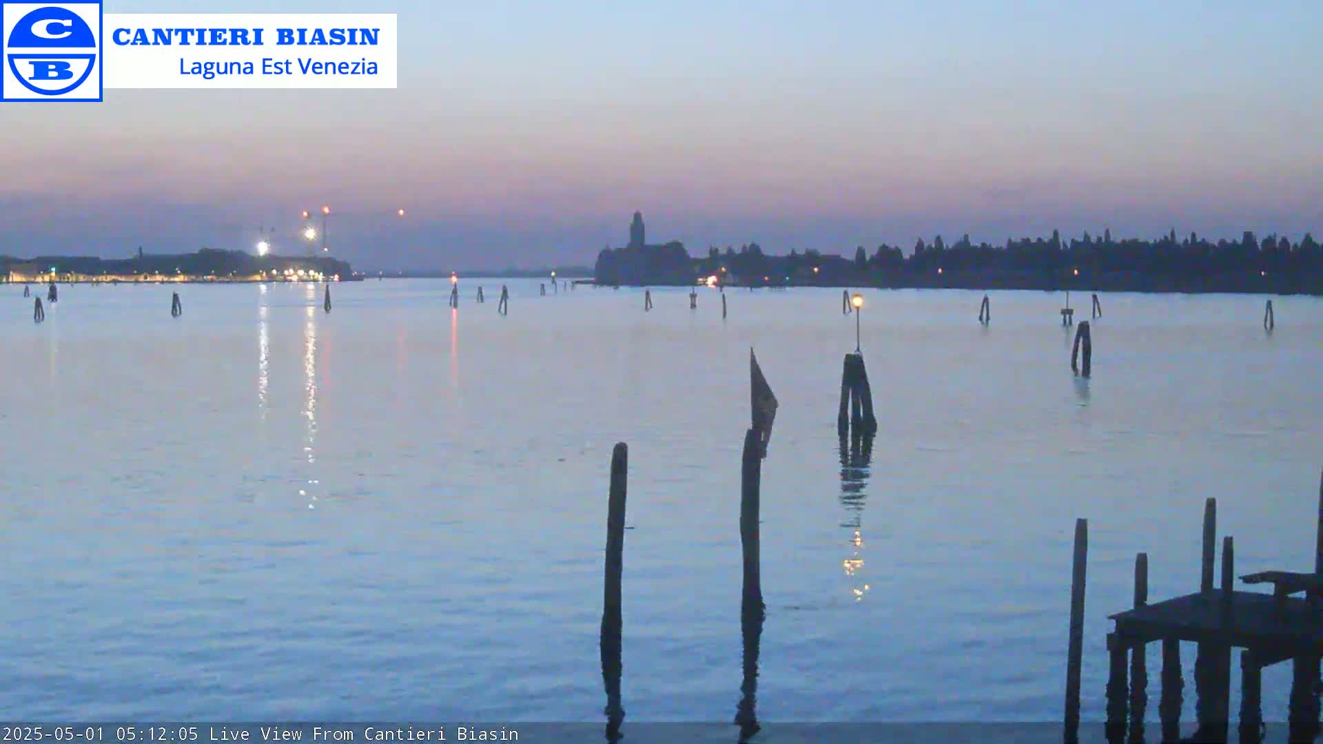 A calm body of water at twilight shows a distant city skyline, illuminated lights, and several wooden posts in the foreground.
