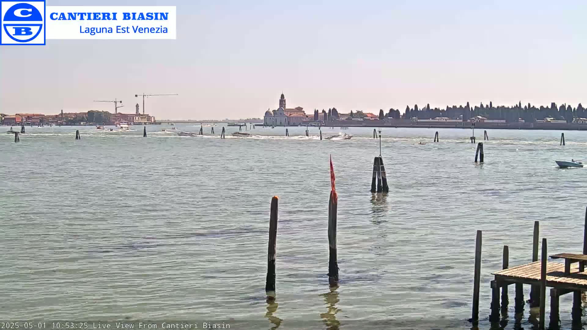 A calm, sunny day shows a body of water with several boats, wooden posts, and a distant island with buildings, including a church tower.