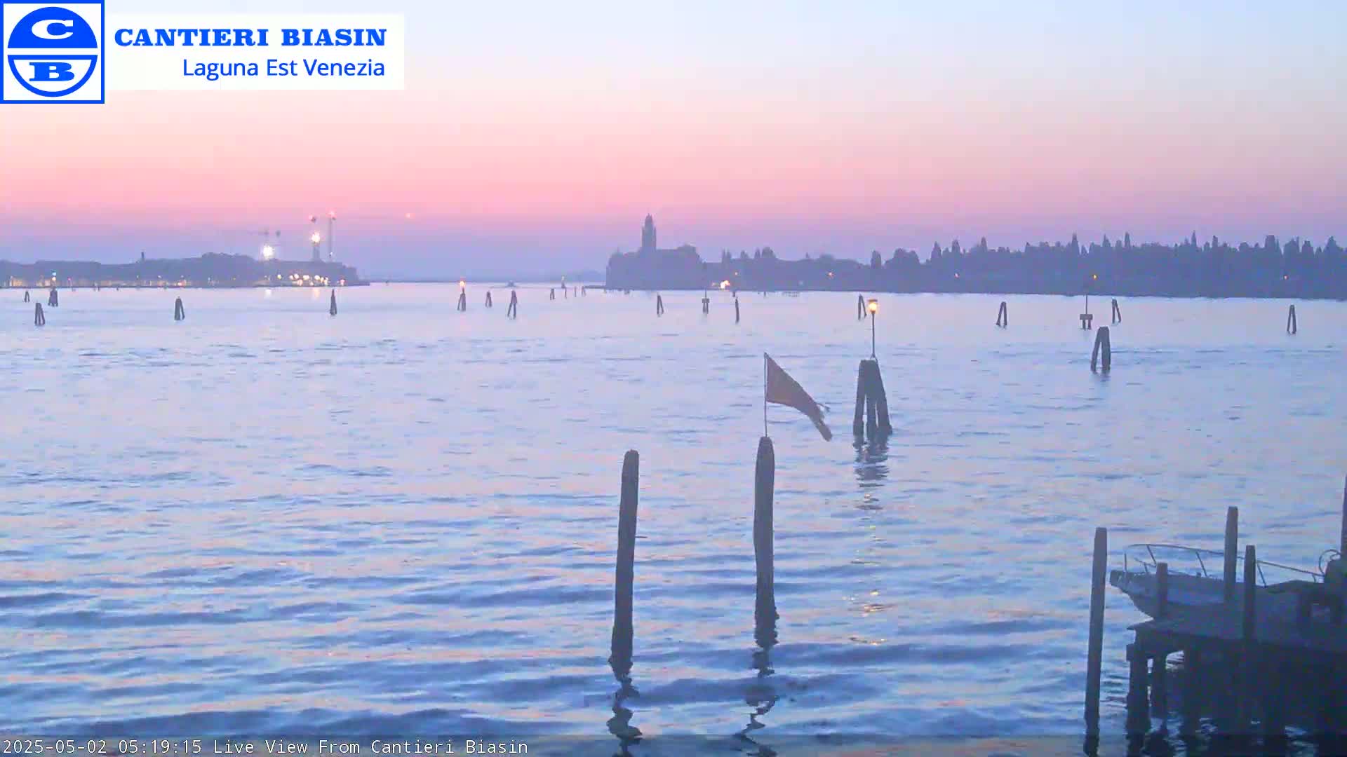 A calm body of water at dusk shows a distant city skyline under a pale pink and purple sky, with several wooden posts and a small boat visible in the foreground.