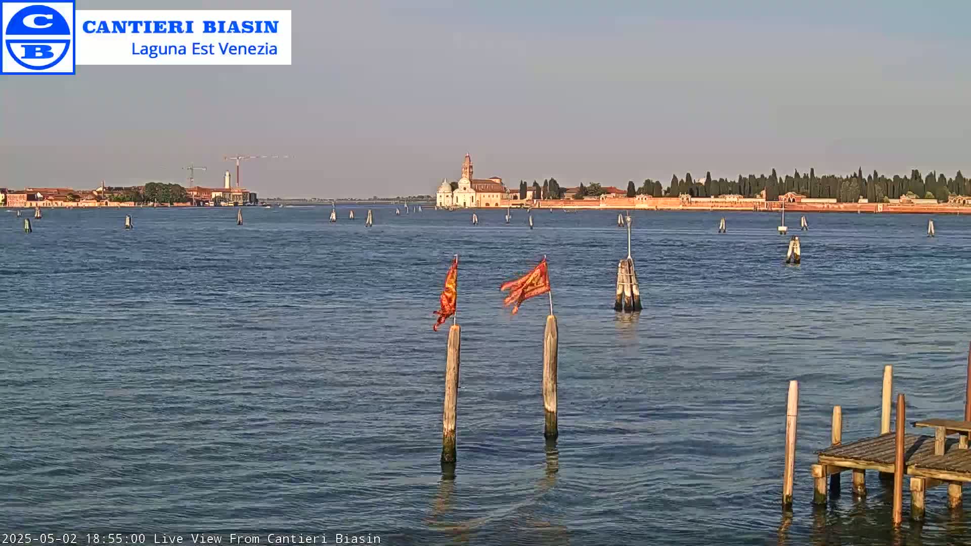 A calm body of water shows a distant island with buildings and trees under a clear sky, two orange flags waving on wooden posts in the foreground.