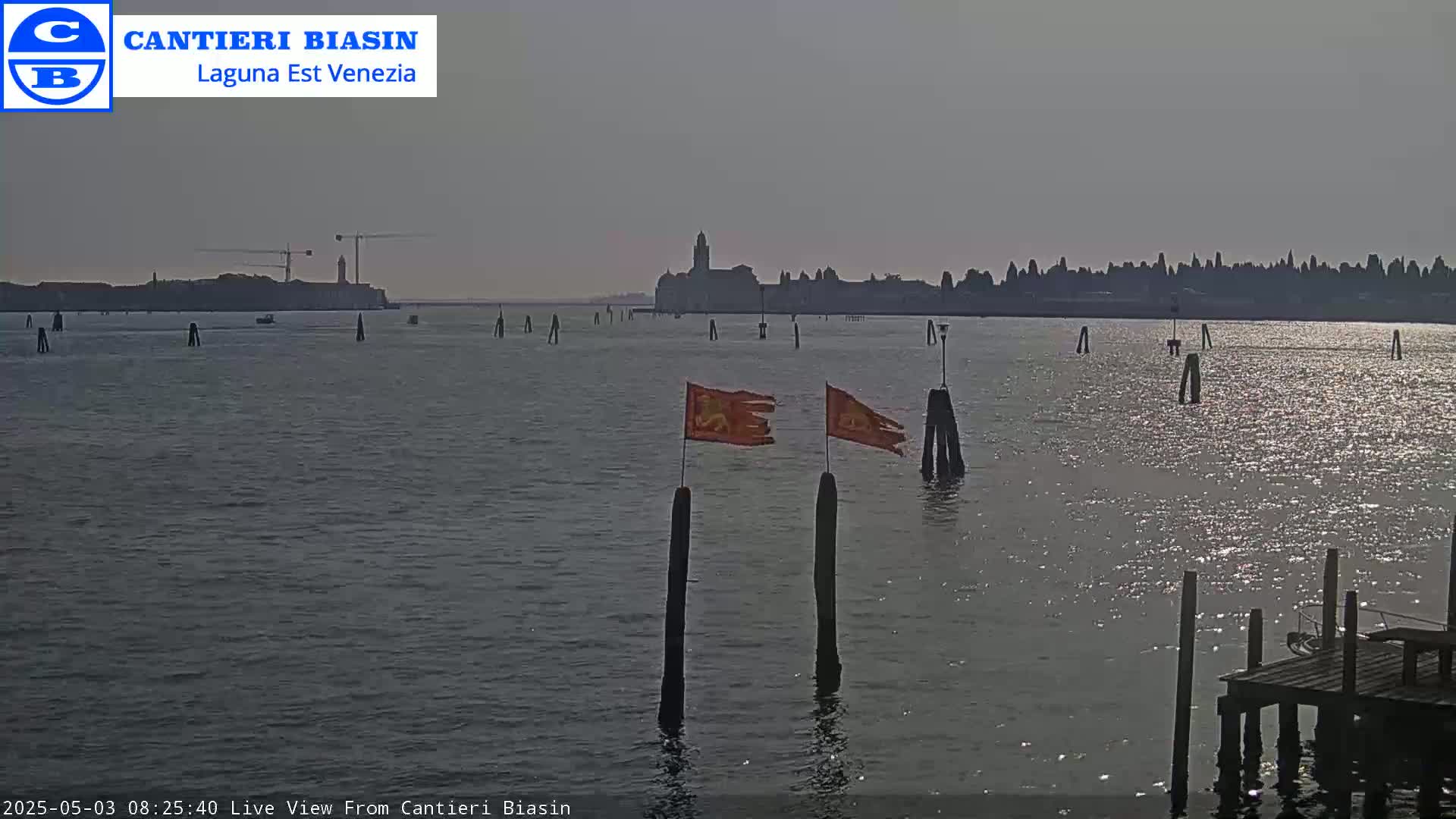 A calm body of water displays two orange flags on posts in the foreground, with a distant cityscape under a hazy sky.