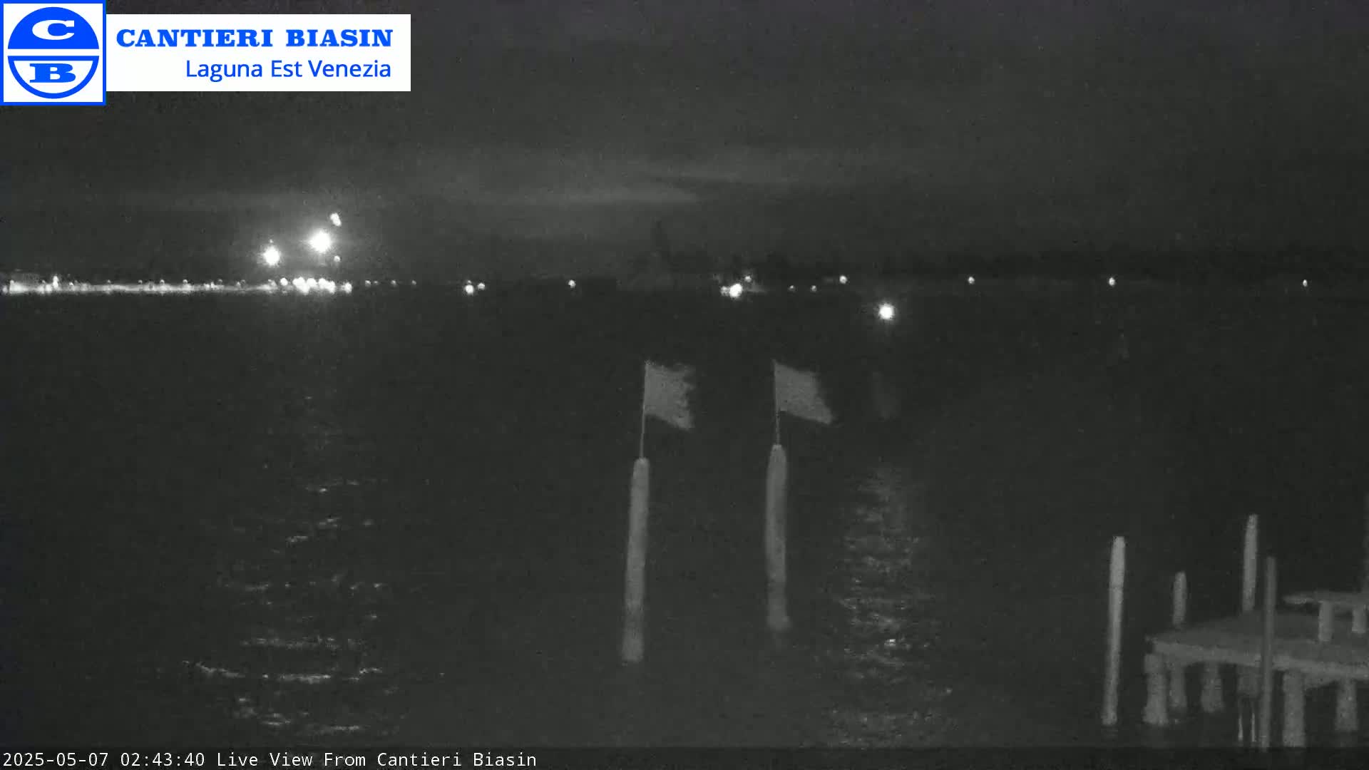 A nighttime view of calm water shows a distant shoreline with lights, two flags on poles in the foreground, and a small dock.