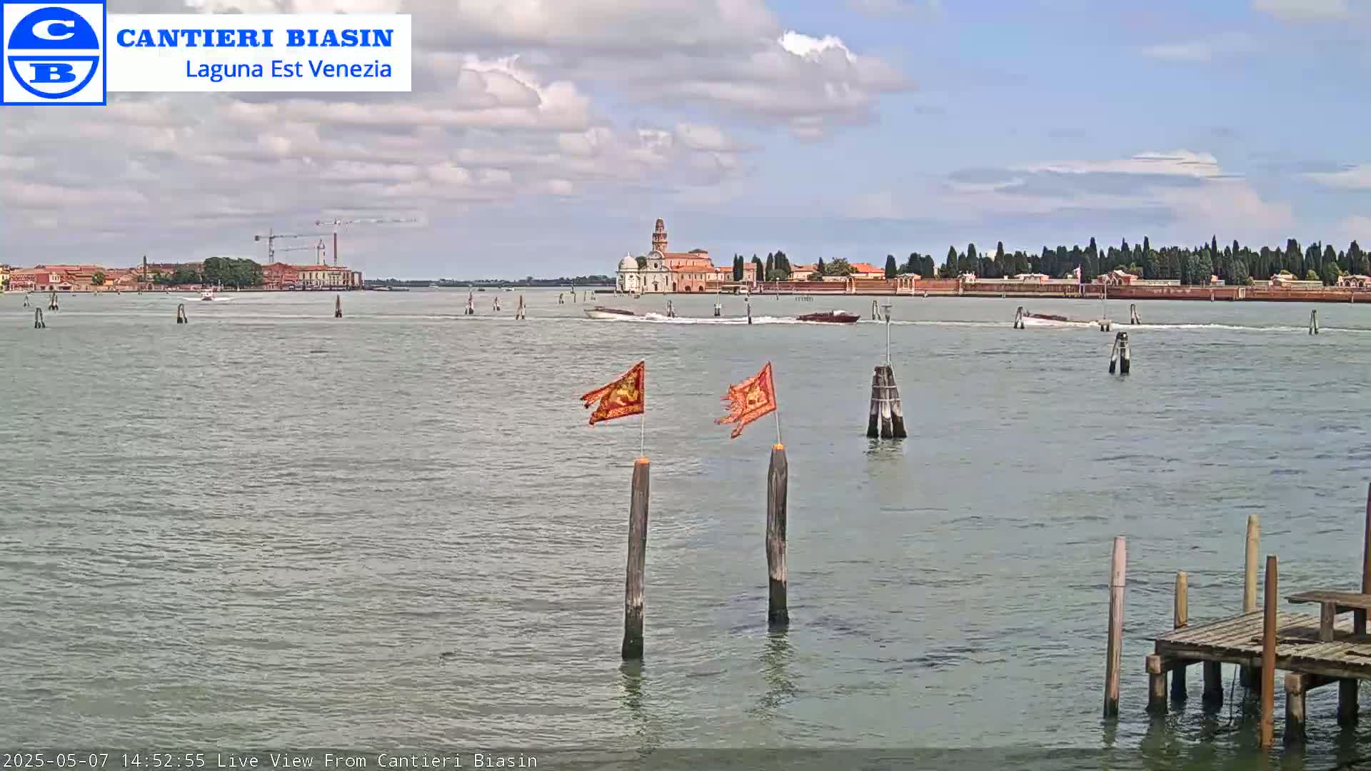 A calm, partly cloudy day shows a view of a Venetian island with buildings and trees across a body of water, featuring two weathered flags on wooden posts in the foreground.