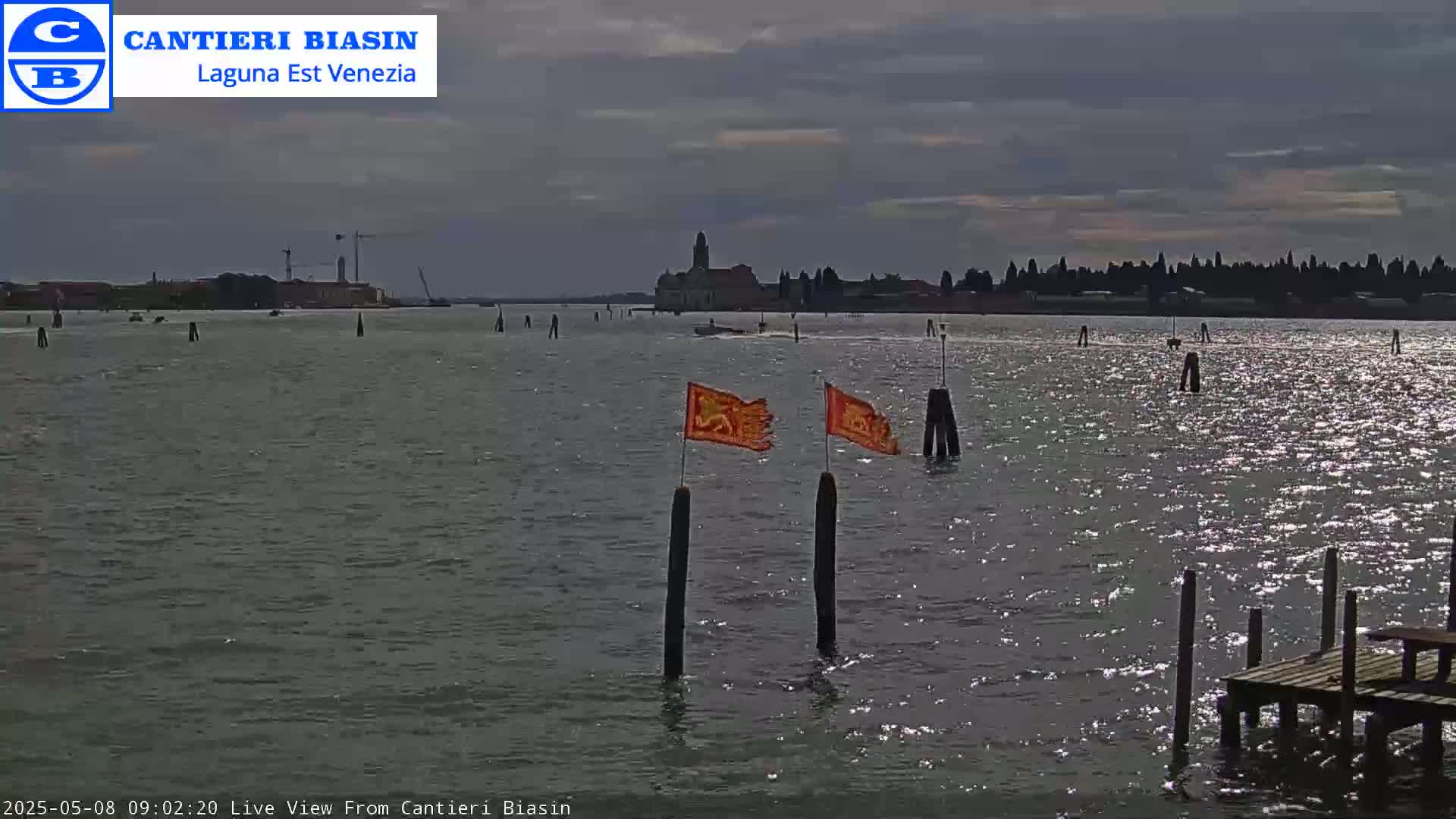 A body of water with a distant cityscape under a cloudy sky, featuring two orange flags on posts in the foreground and a small dock.
