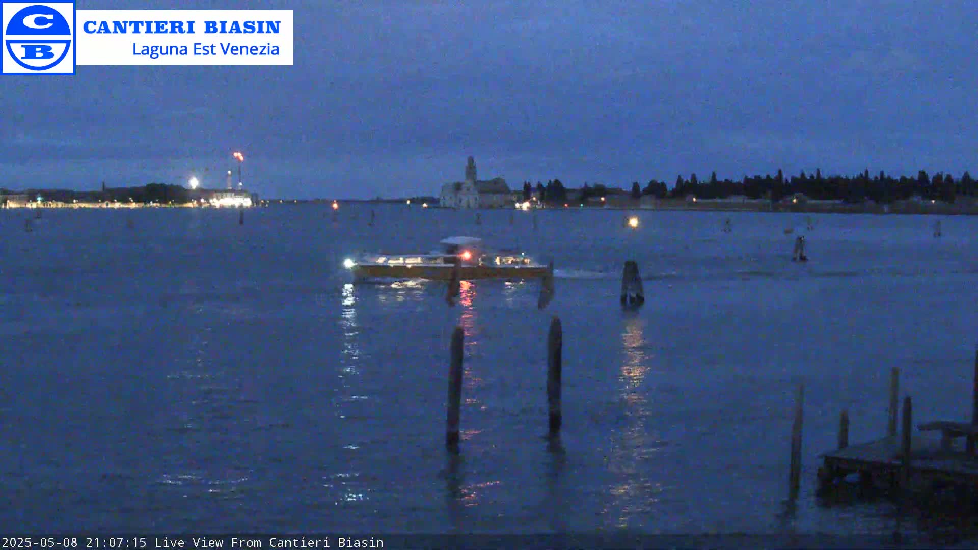 A boat travels across dark blue water at twilight, past several posts in the water and a distant shoreline with buildings.