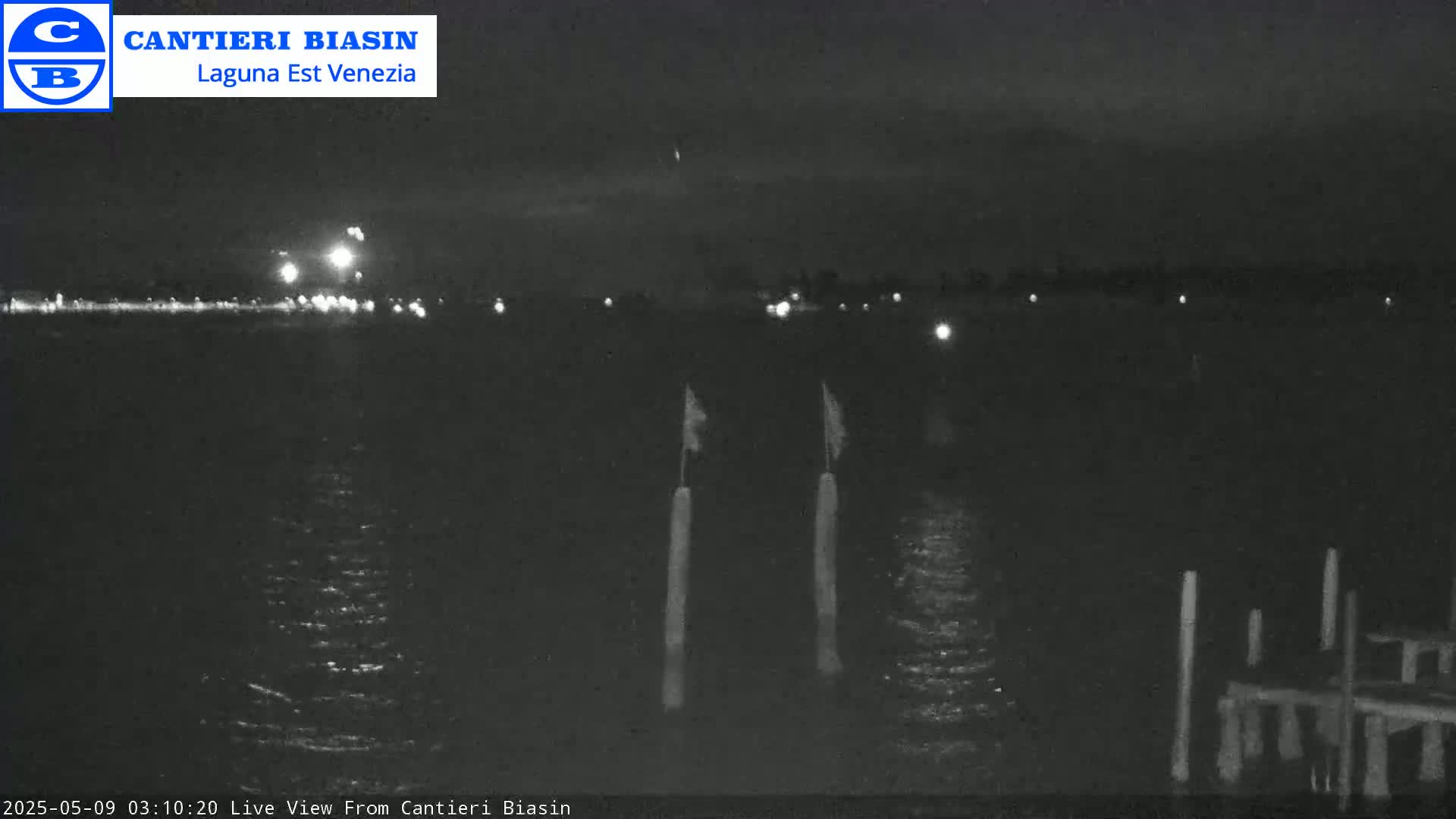 A nighttime view of a dark body of water with lights visible in the distance and two tall posts with small flags on them in the foreground.