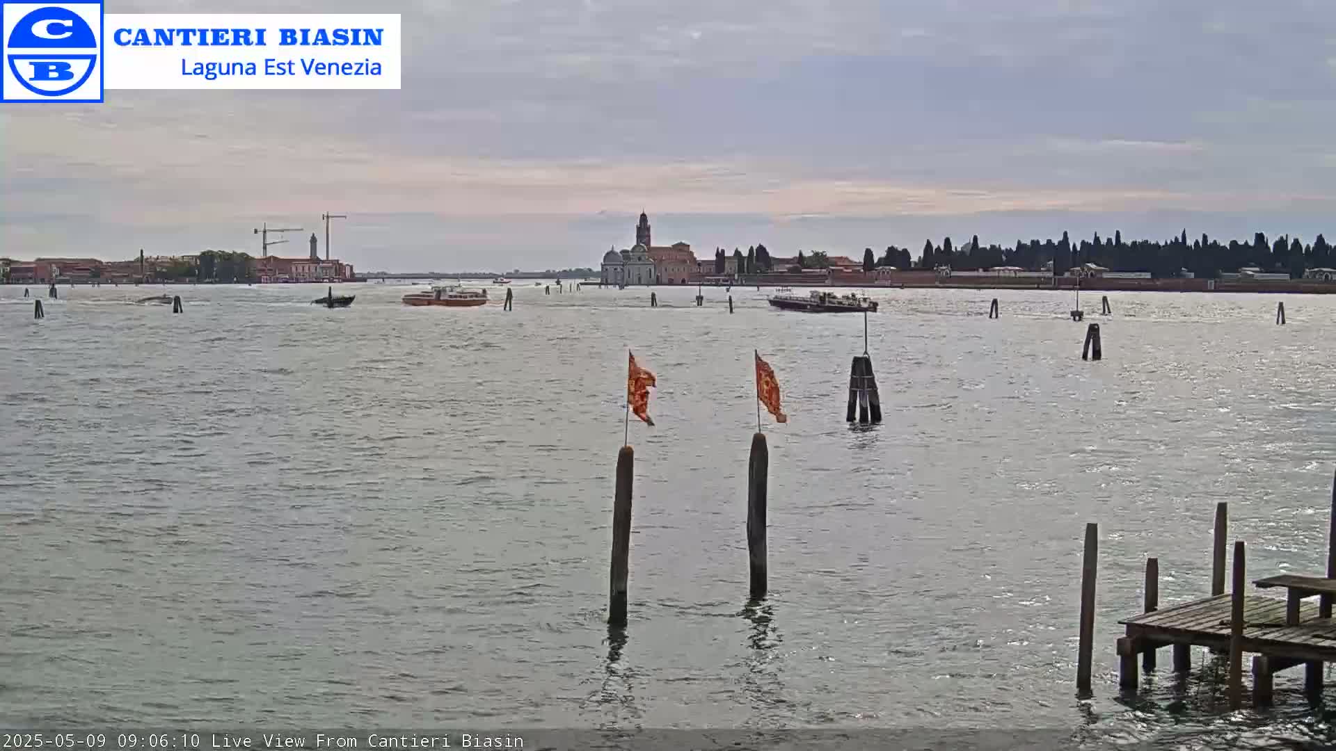 A calm, overcast day shows a view of a waterway with boats, a distant island with buildings, and wooden posts with small orange flags in the foreground.