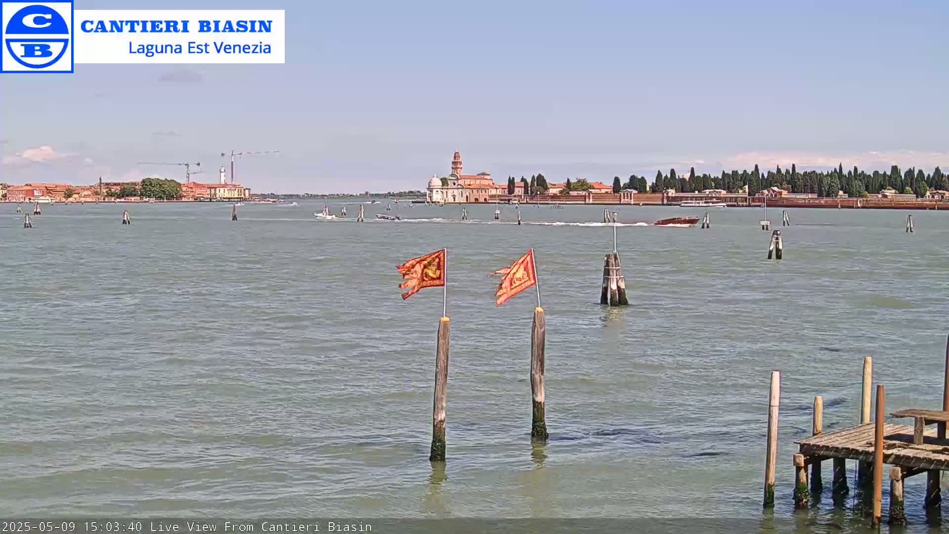 A calm body of water shows a distant island with buildings under a clear sky, featuring two orange flags on wooden posts in the foreground and a small wooden dock.