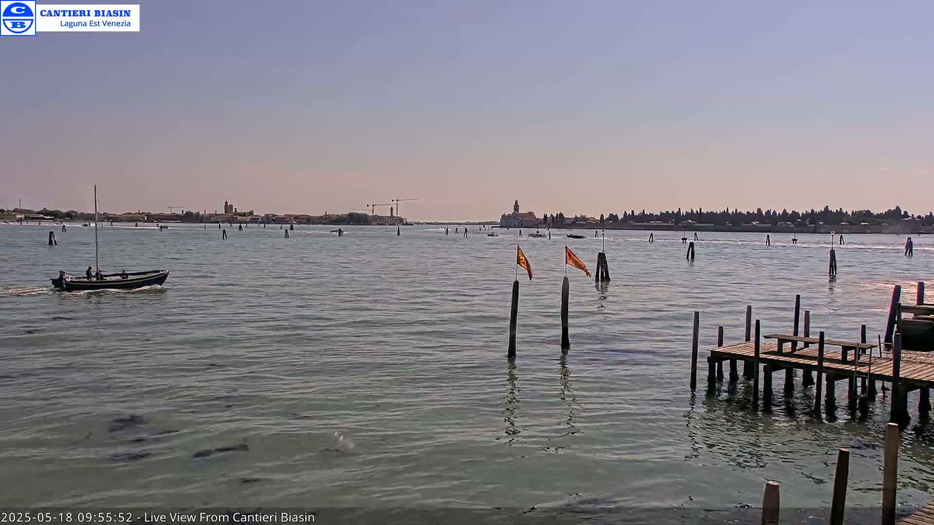 A calm body of water shows a small boat, a distant shoreline, wooden pilings with a few flags, and a simple wooden dock under a clear, sunny sky.